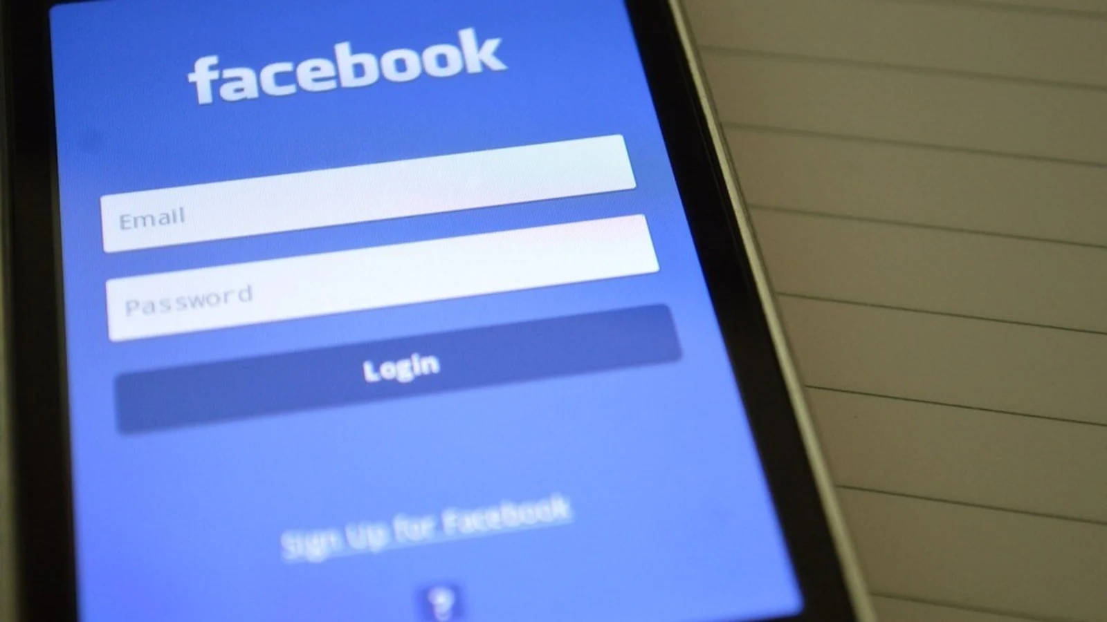 手機與記事本登入 Facebook 壁紙 — 智能手機特寫，顯示Facebook登入畫面，包含電郵和密碼欄位，背景為橫線筆記本。