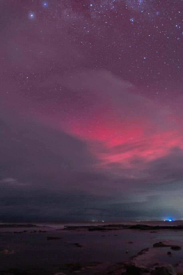  壁紙 — 繁星點點的夜空，充滿活力的粉紅色極光在黑暗的沿海景觀上閃耀，遠處有燈光。