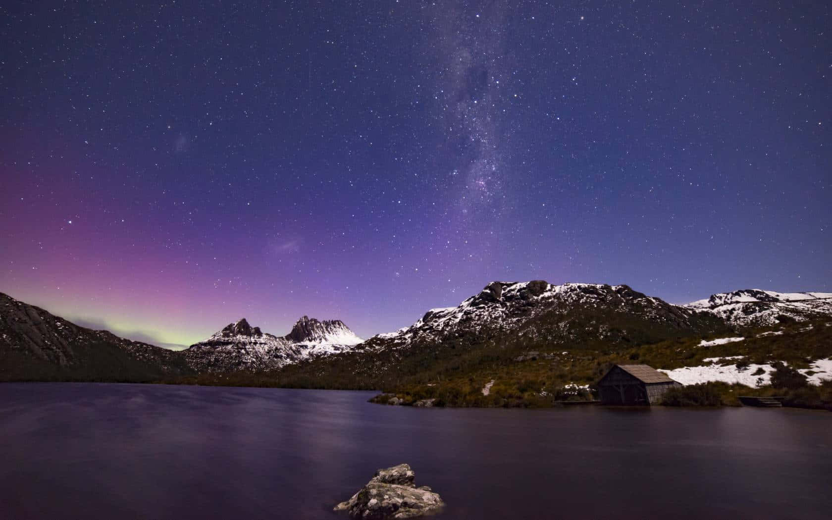  壁紙 — 繁星點點的夜空，絢麗的極光拱形橫跨在積雪的山脈和寧靜的湖泊上，湖邊有一座小木屋。