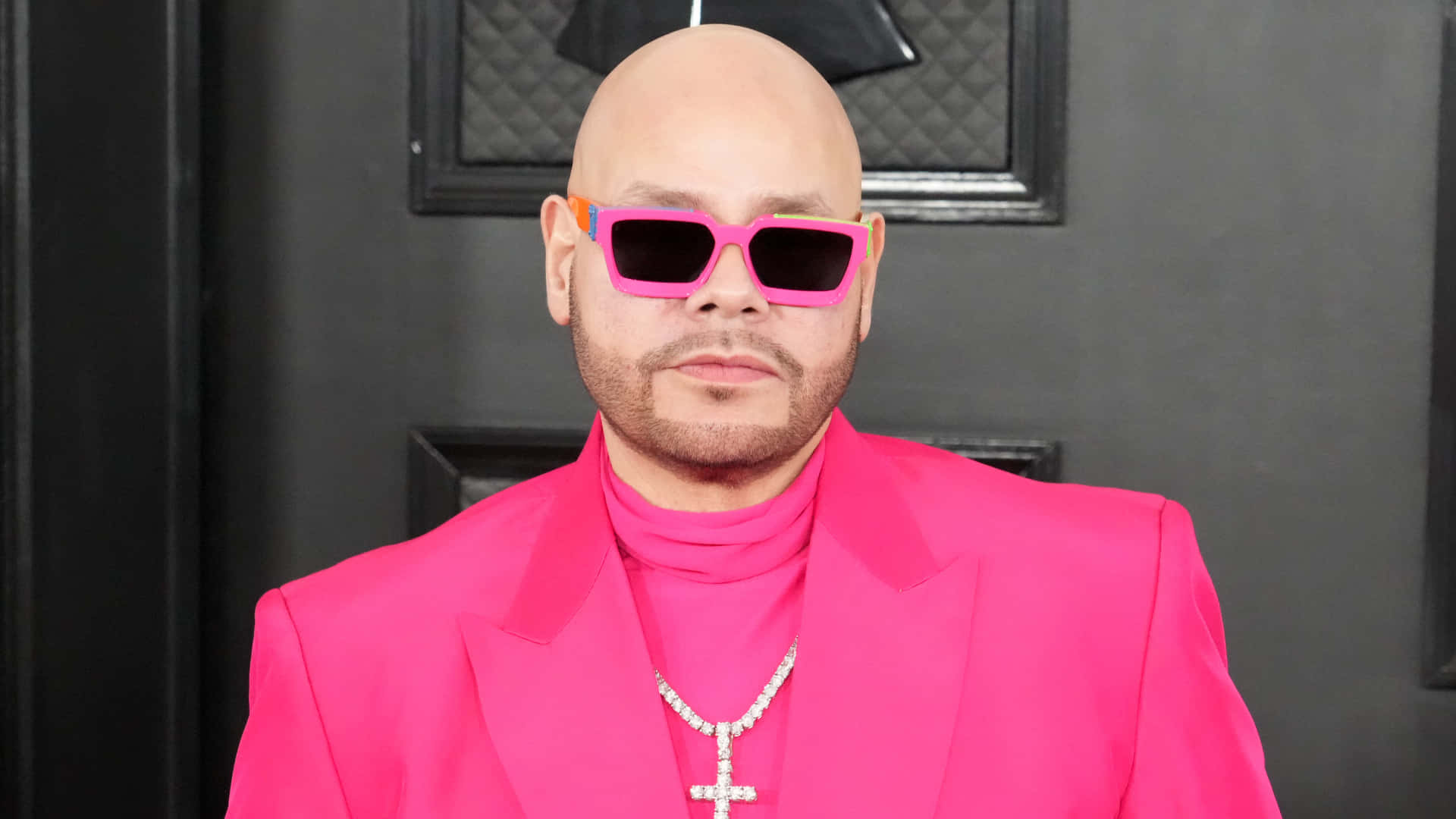 Fat Joe 身著粉紅服裝出席活動 壁紙 — 一名光頭男子穿著大膽的粉紅色西裝和相配的太陽眼鏡，在深色紋理牆壁前自信地擺姿勢。