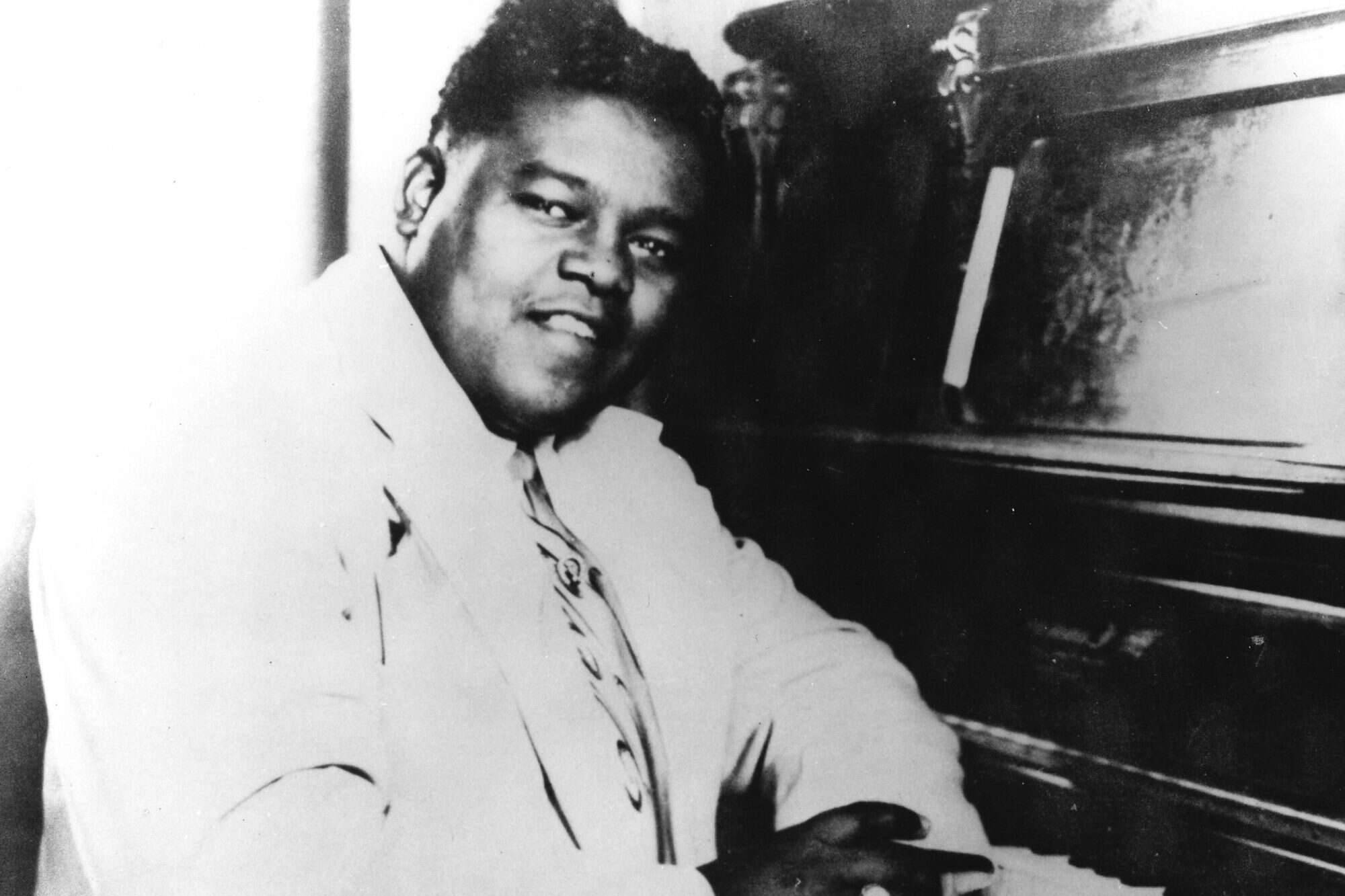 Fats Domino — 搖滾樂傳奇 壁紙 — 一位微笑的黑人男士穿著淺色西裝坐在鋼琴前，在復古黑白照片中溫暖地看著鏡頭。