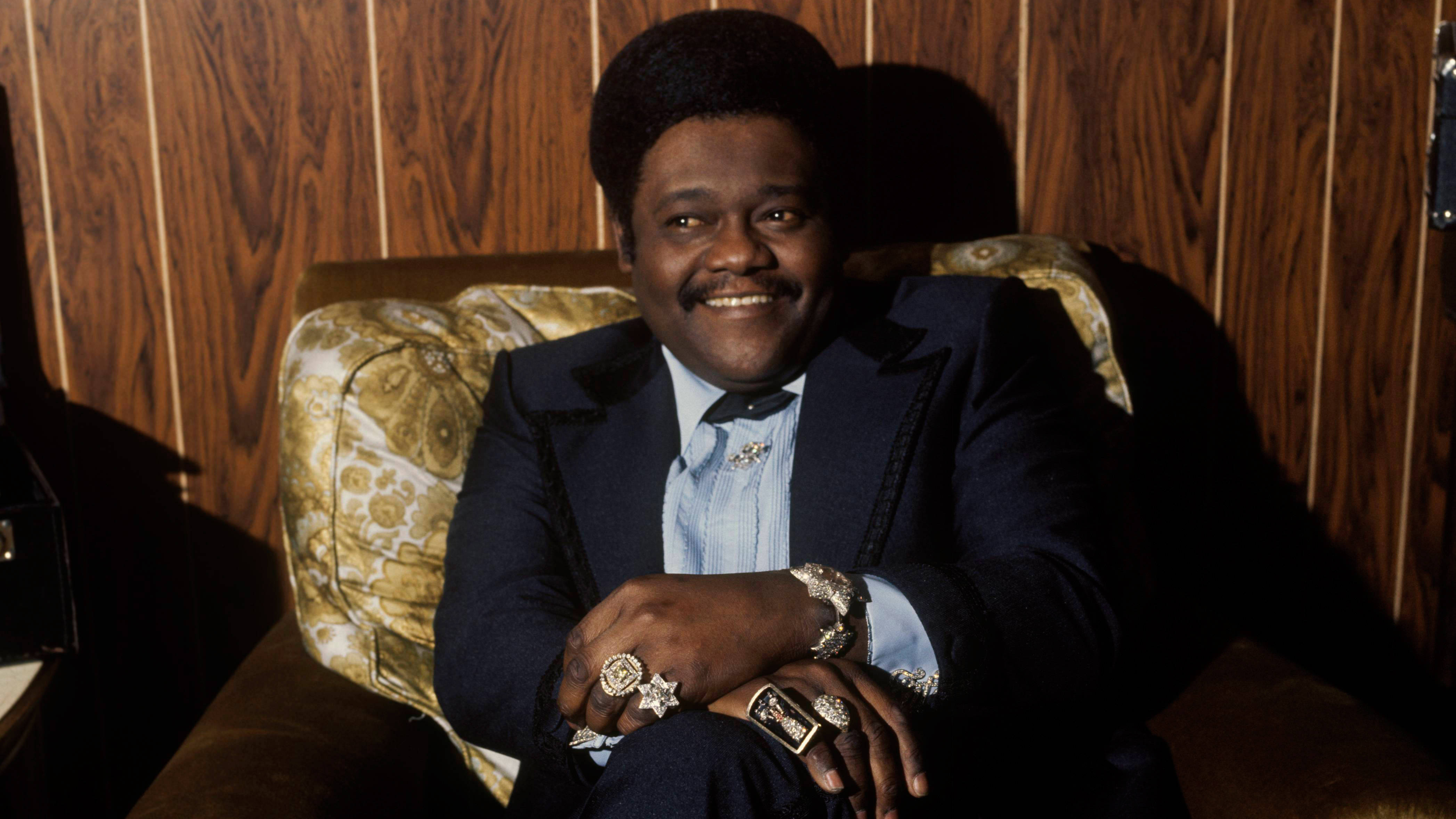 Fats Domino 摇滾先驅 壁紙 — 一個微笑的男人穿著深色西裝和淺藍色襯衫，自信地坐在有圖案的椅子上，背景是木質牆板。