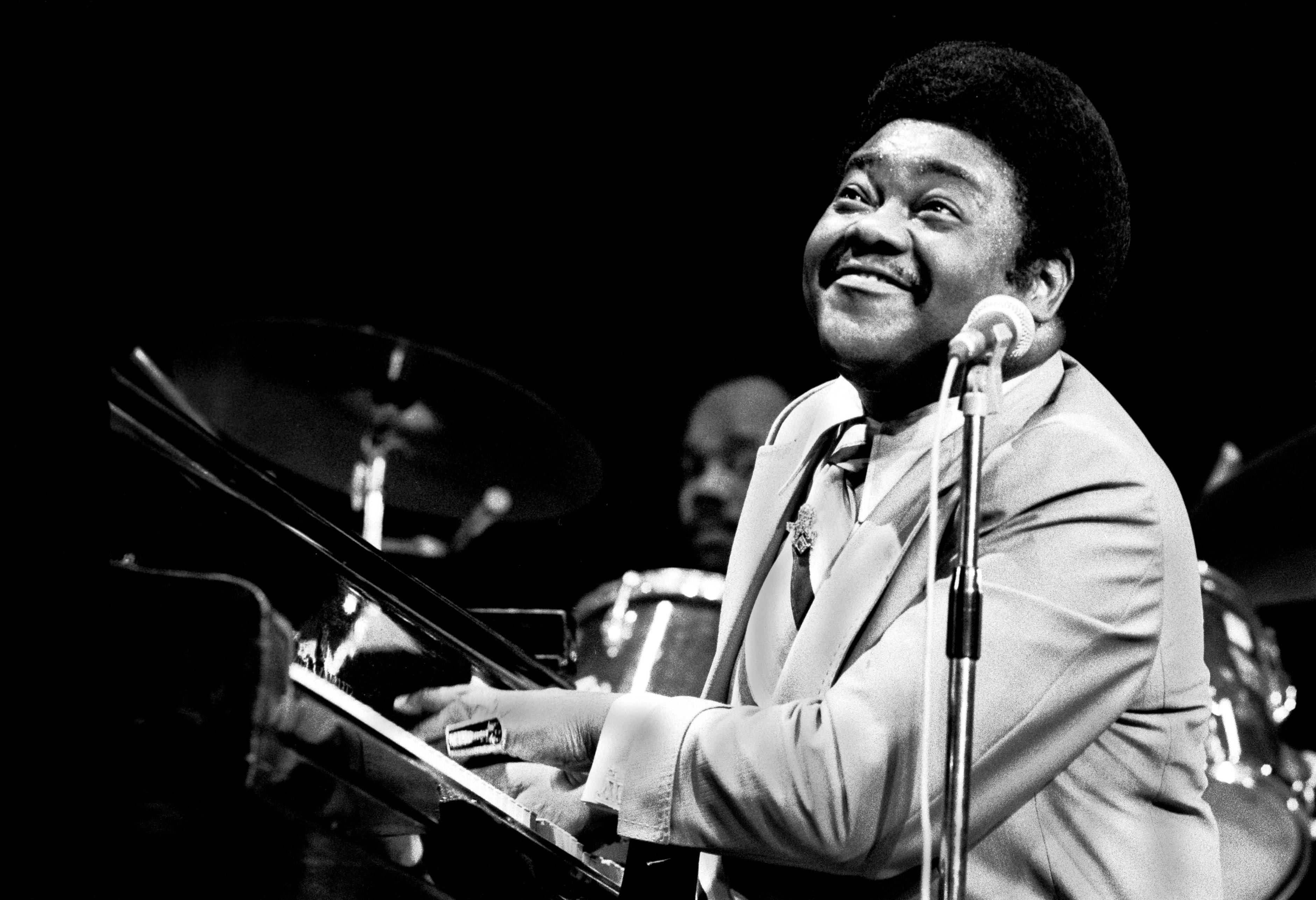 Fats Domino 經典舊照 壁紙 — 一位快樂的爵士鋼琴家在舞台燈光下彈奏三角鋼琴，身後有鼓手，溫暖地微笑。