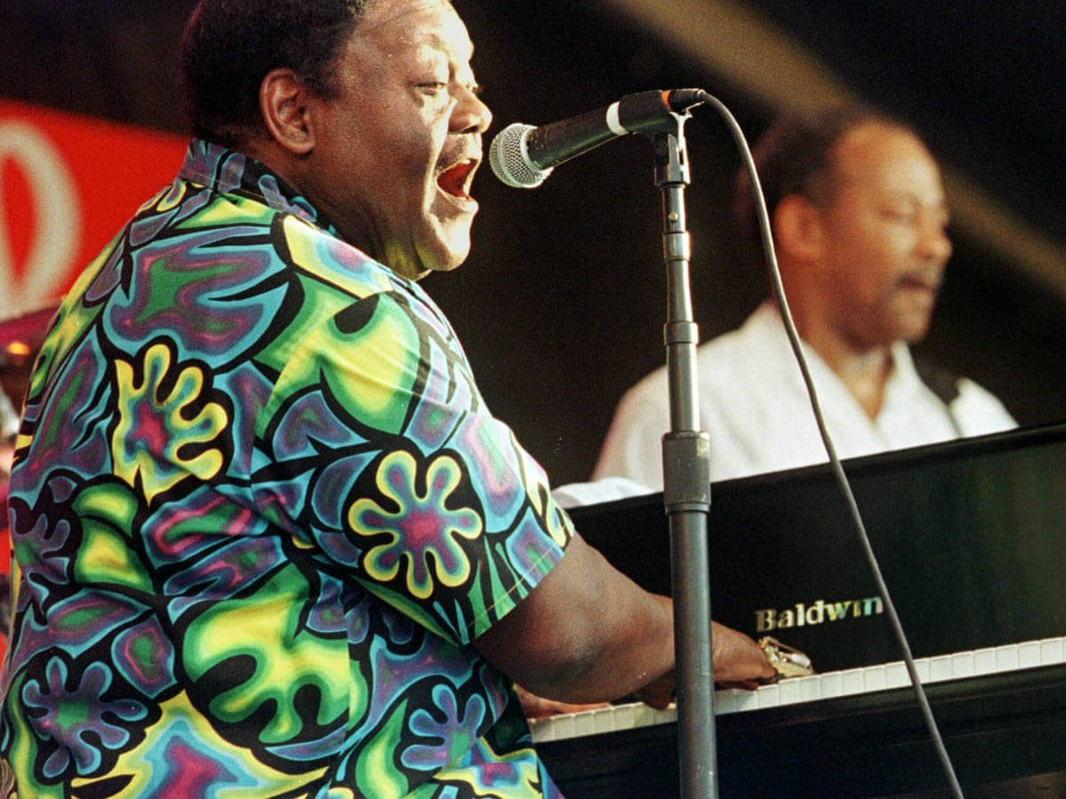 Fats Domino 美國鋼琴家歌手 壁紙 — 一位充滿熱情的音樂家穿著鮮豔的花襯衫，在舞台上對著麥克風唱歌，同時彈奏鮑德溫鋼琴。