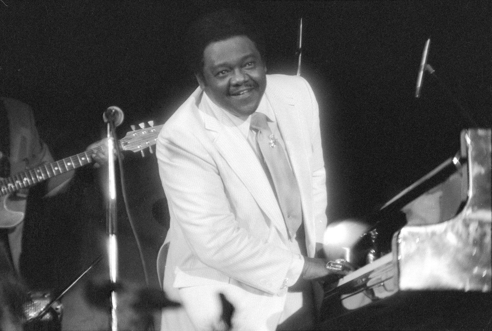 Fats Domino 美國鋼琴家 壁紙 — 一位微笑的白西裝男子在舞台上明亮的聚光燈下彈鋼琴，背景有一位結他手。