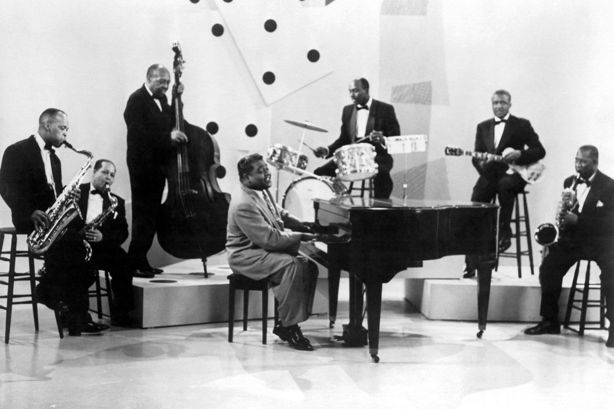 Fats Domino 與他的樂隊 壁紙 — 一張黑白照片，爵士樂隊在帶有幾何牆壁圖案的現代主義舞台上表演。