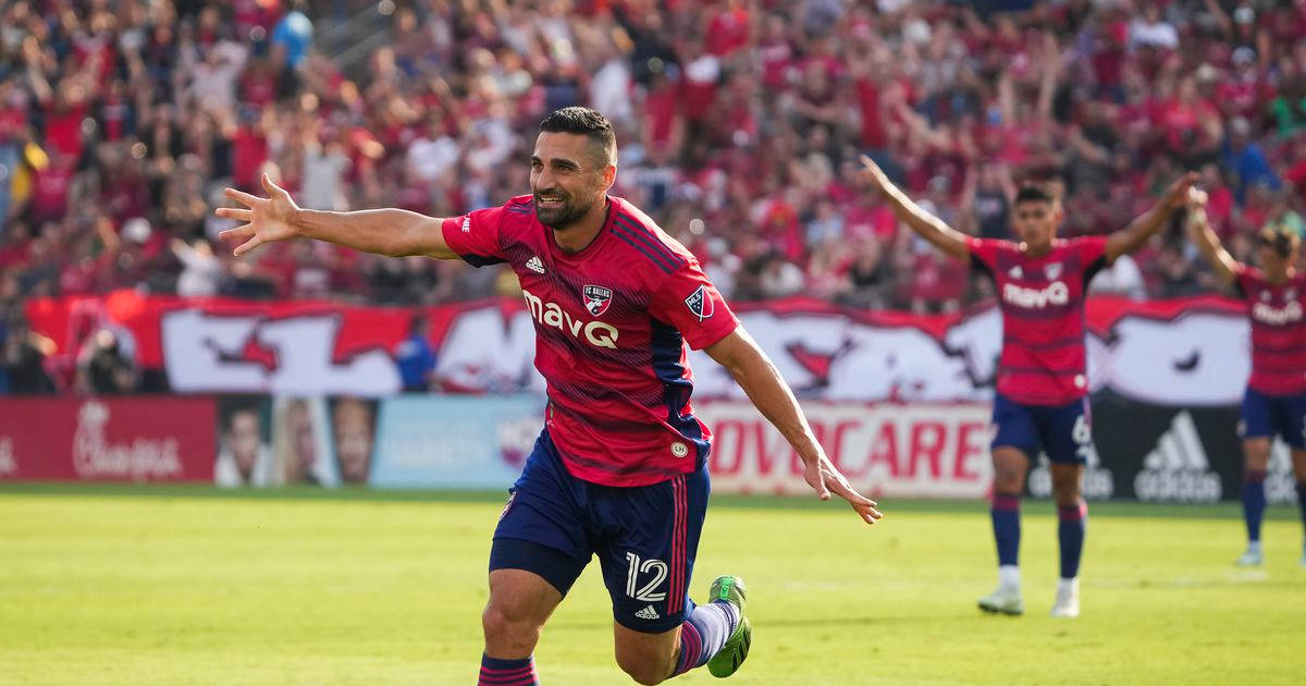 FC達拉斯職業運動員塞巴斯蒂安·萊特蓋特 壁紙 — 一名身穿紅色球衣的足球運動員在陽光明媚的綠色球場上張開雙臂慶祝入球，背景中隊友和球迷在歡呼。