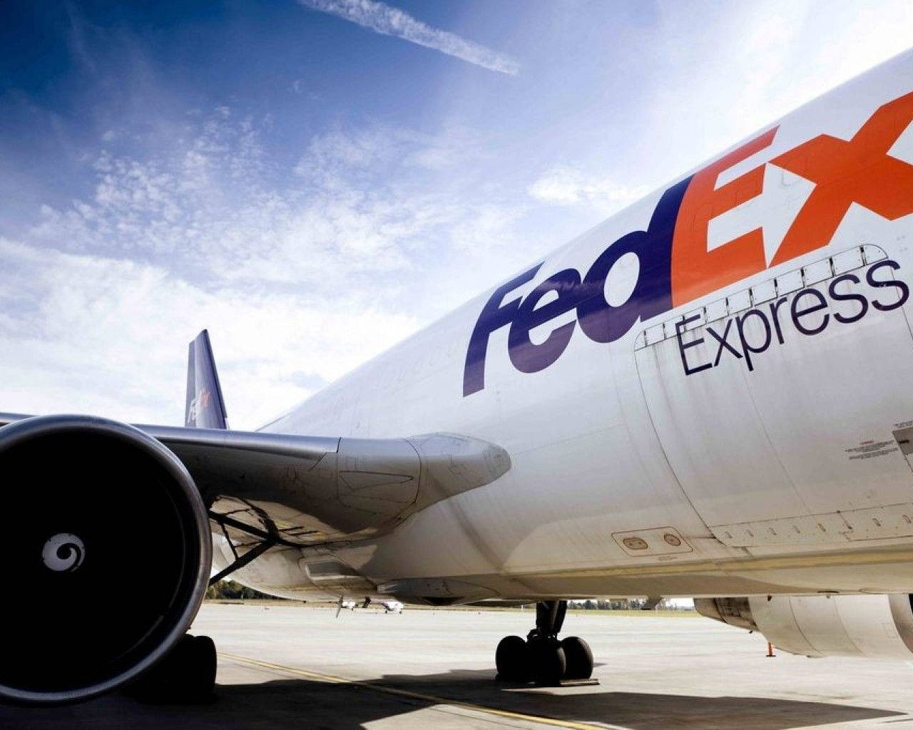 FedEx Express飛機側面特寫 壁紙 — 一個低角度拍攝的FedEx Express貨機，在陽光明媚的停機坪上，藍天白雲。
