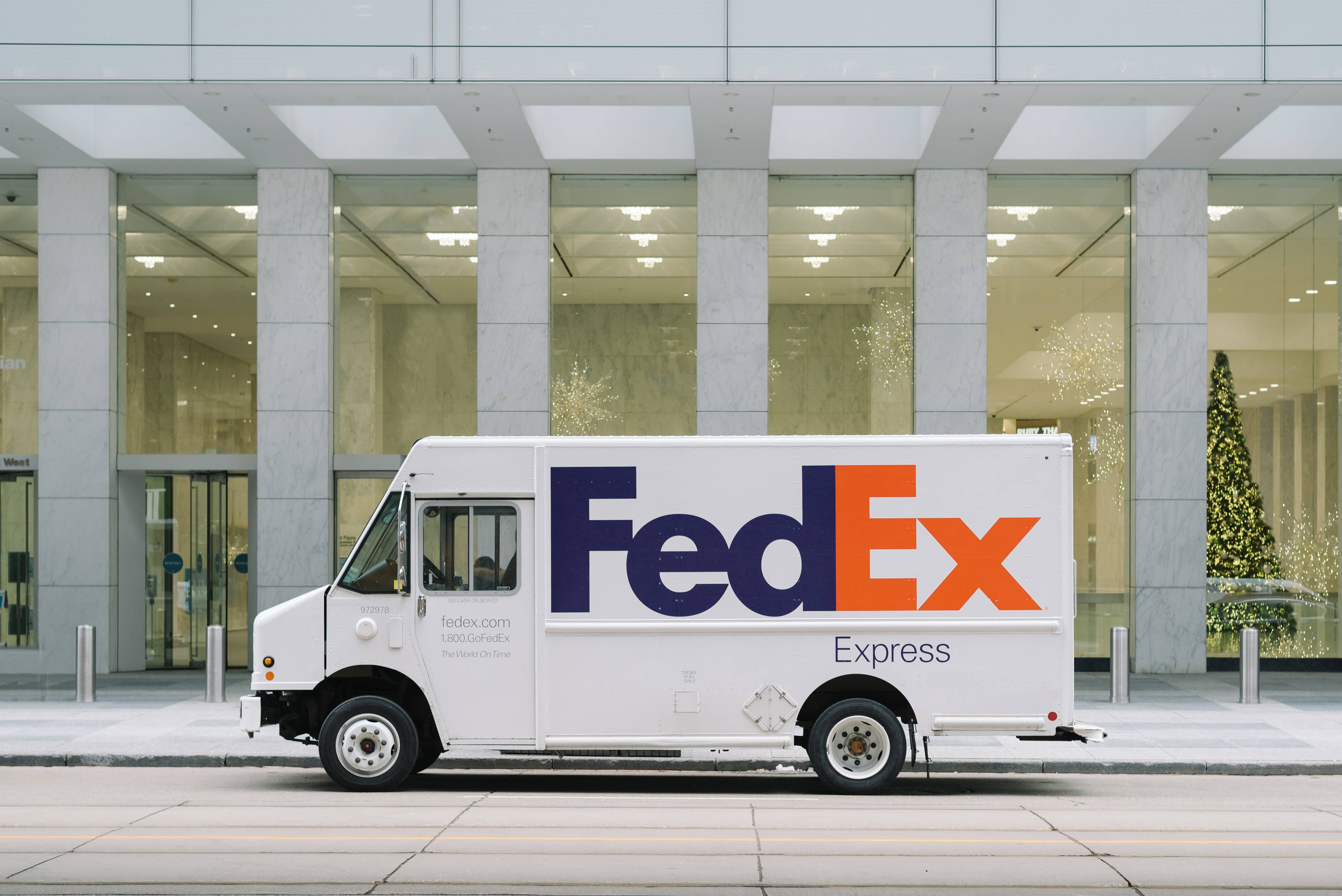FedEx貨運卡車 壁紙 — 一輛白色FedEx Express送貨車停在一棟現代玻璃幕牆建築前，建築內可見一棵亮燈的聖誕樹。