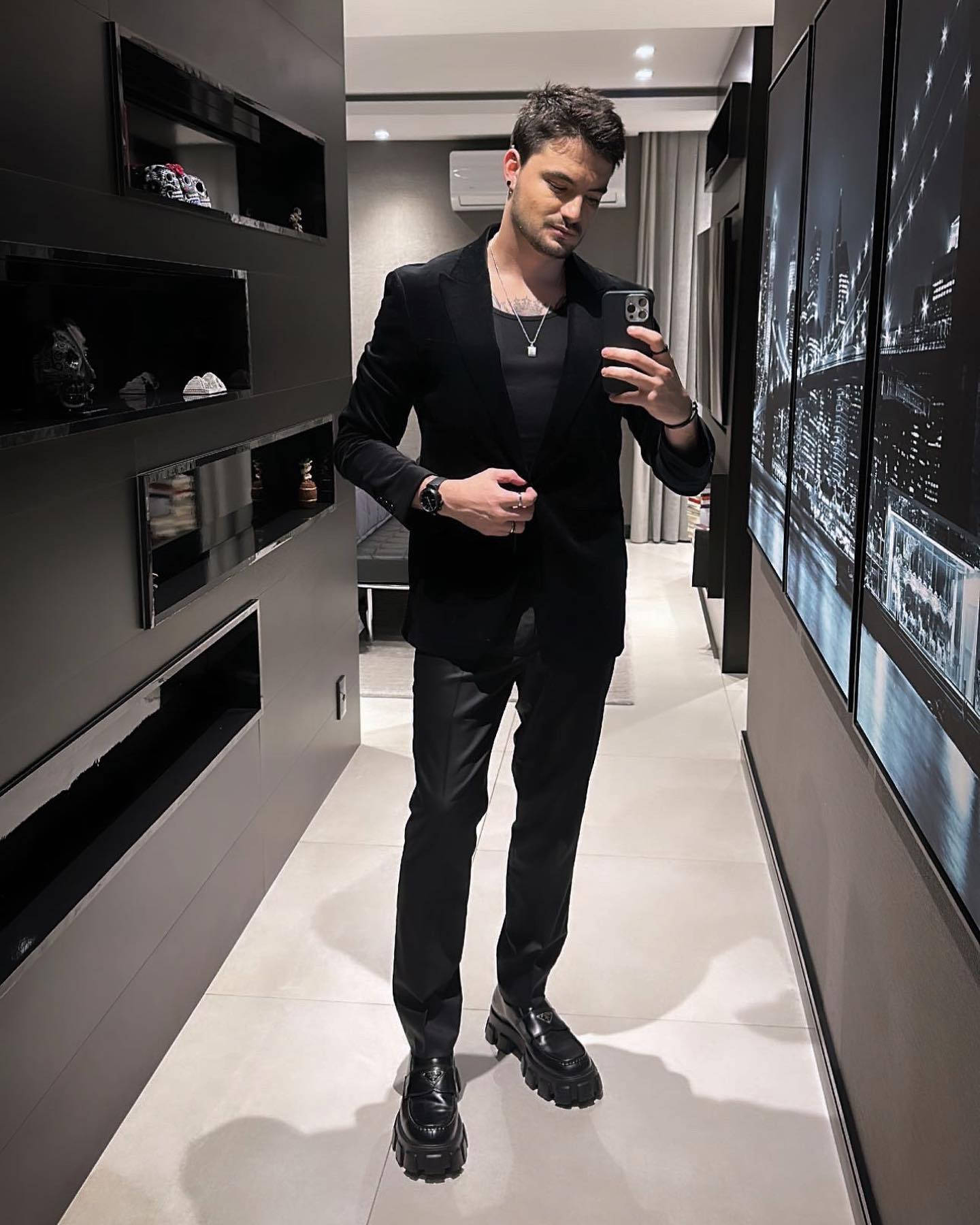 身穿黑色西裝和靴子的男子，在現代走廊裡對著鏡子自拍，牆上掛著城市景觀藝術品。