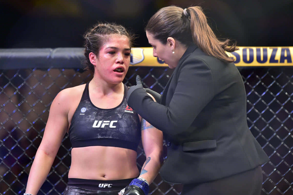  壁紙 — 一位身穿UFC上衣的女性MMA拳手站在八角籠內，教練在休息時檢查她的肩膀。