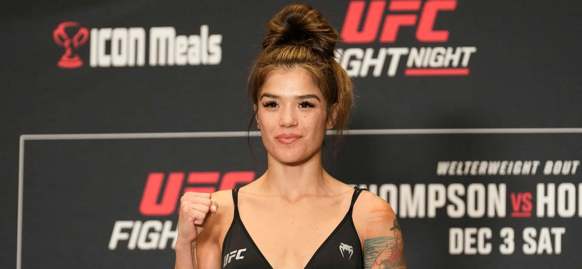  壁紙 — 一位女性UFC拳手自信地擺姿勢，身穿黑色運動內衣，舉起拳頭，背景是品牌活動背景。