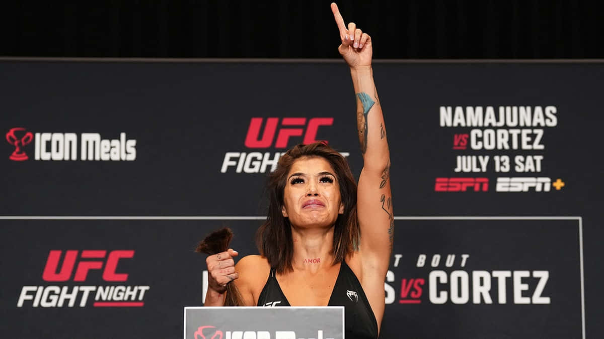  壁紙 — 一名女性UFC拳手在稱重活動中勝利地舉起食指，背後有「Namajunas vs Cortez」的品牌標誌。