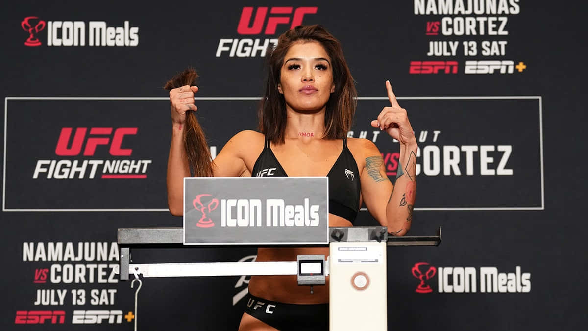  壁紙 — 一位女性UFC拳手在稱重活動中自信地站在體重秤上，身後有宣傳橫幅。