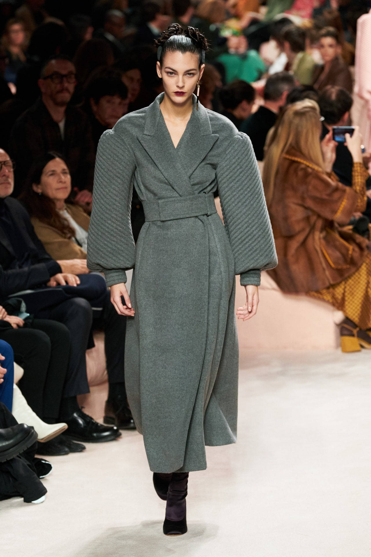 Fendi 厚實浴袍 壁紙 — 一位模特兒穿著結構感灰色外套，帶有寬鬆袖子和腰帶，在伸展台上行走，周圍坐著觀眾。