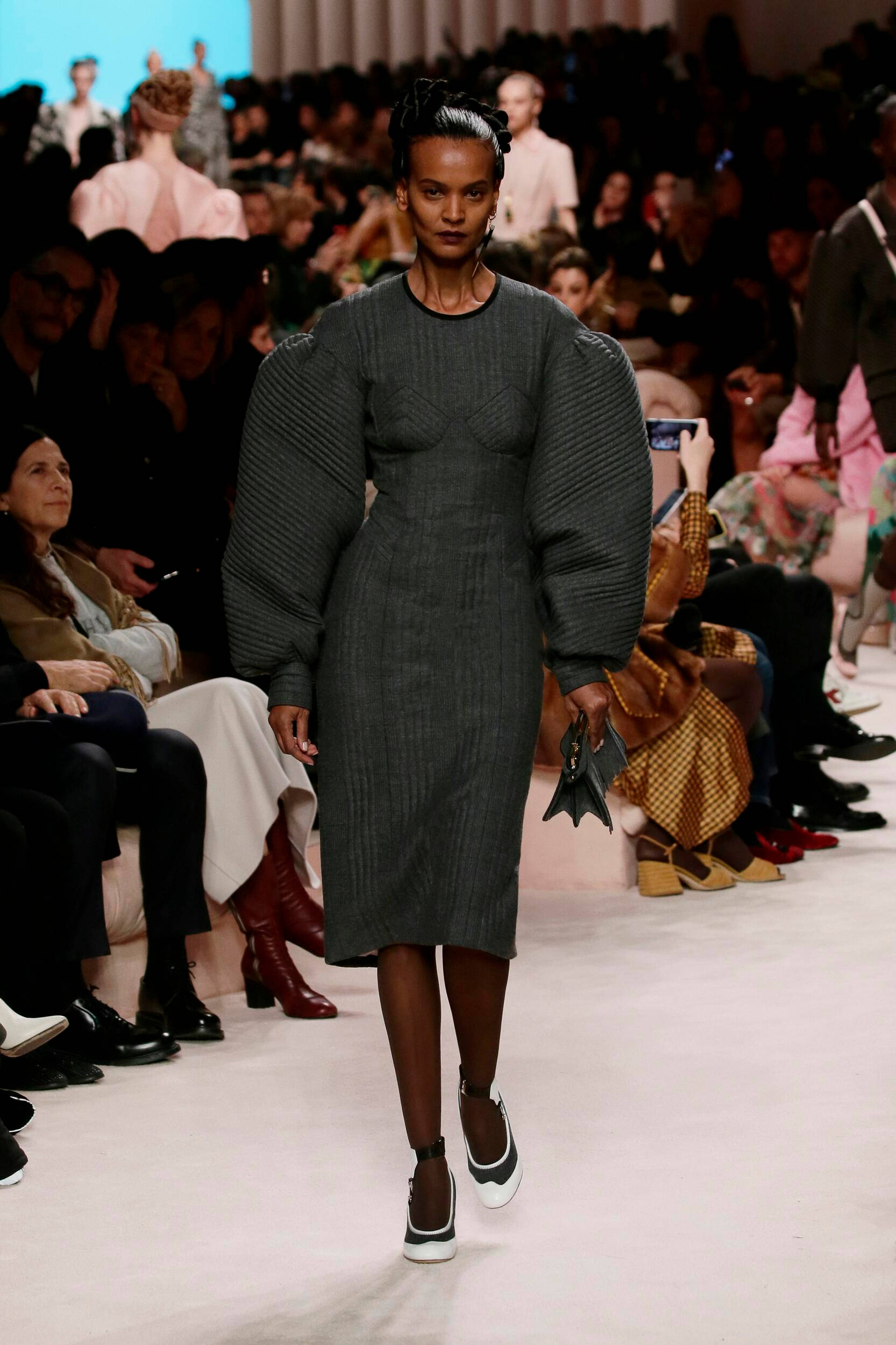 Fendi 深綠色禮服 壁紙 — 一位模特兒自信地走在天橋上，身穿結構感灰色連衣裙，搭配戲劇性的泡泡袖和黑白運動鞋。