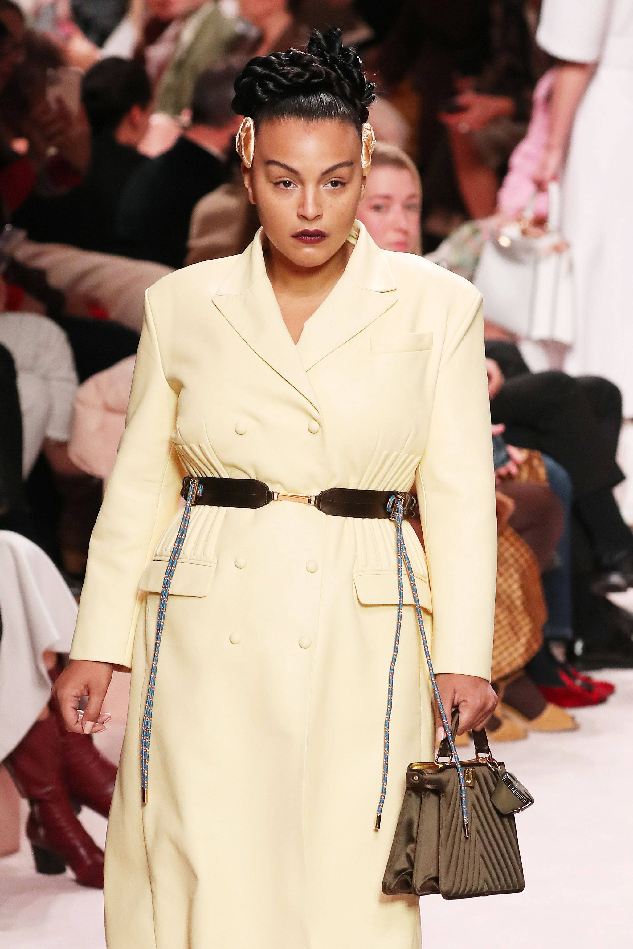 Fendi 米色大衣 壁紙 — 模特兒自信地走在天橋上，身穿淡黃色雙排扣外套，配有寬腰帶，並手持結構感十足的綠色手袋。