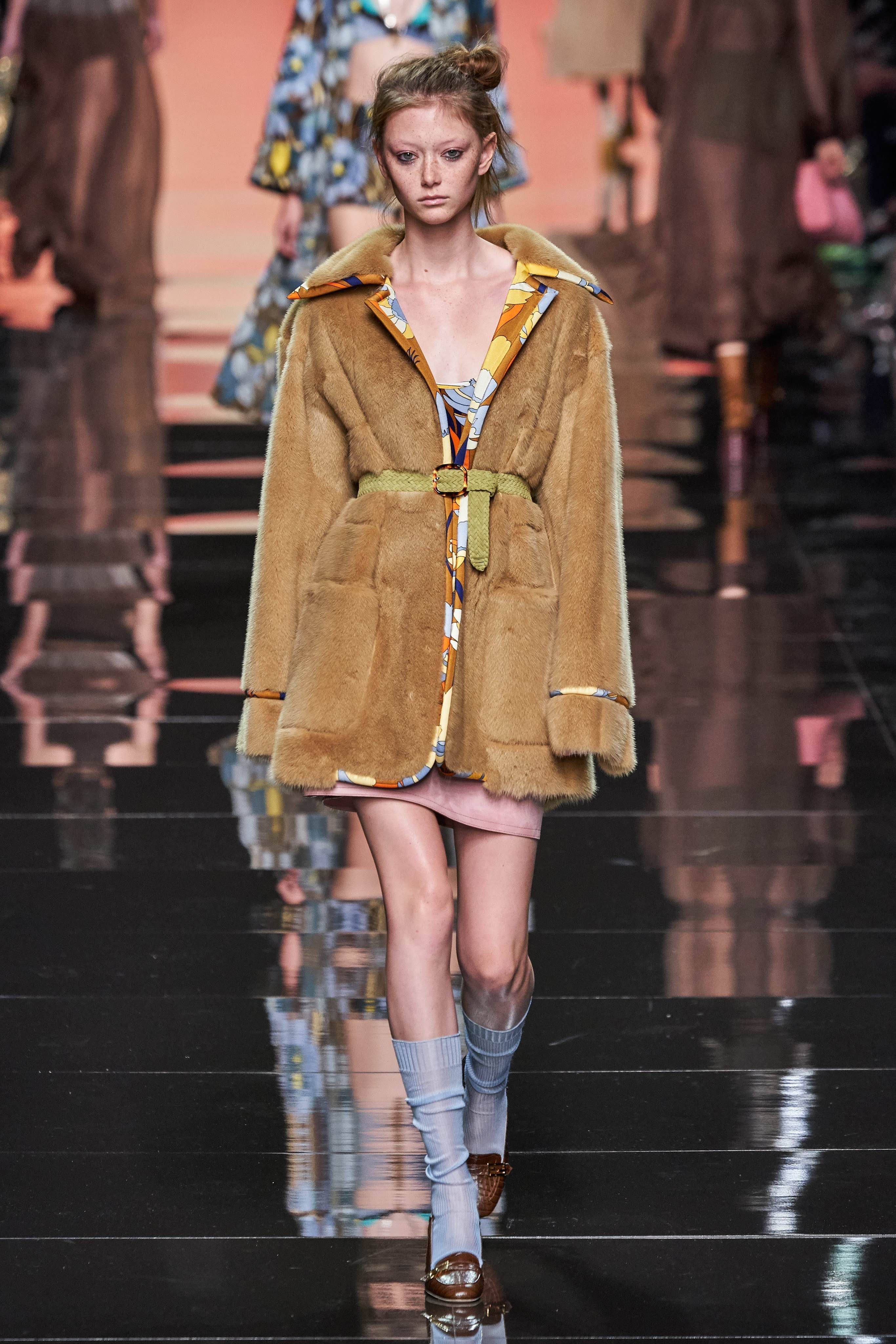 Fendi 絨毛大衣 壁紙 — 一位模特兒自信地走在光亮的時裝天橋上，身穿帶有圖案飾邊的棕褐色毛皮大衣和淺藍色襪子。