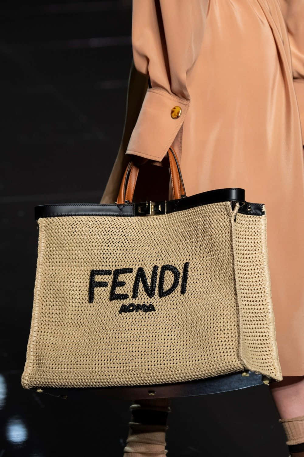 FENDI的奢華美學 壁紙 — 模特兒手持一款編織FENDI ROMA手提袋，配以黑色皮革飾邊和手柄，背景為昏暗的時裝天橋。