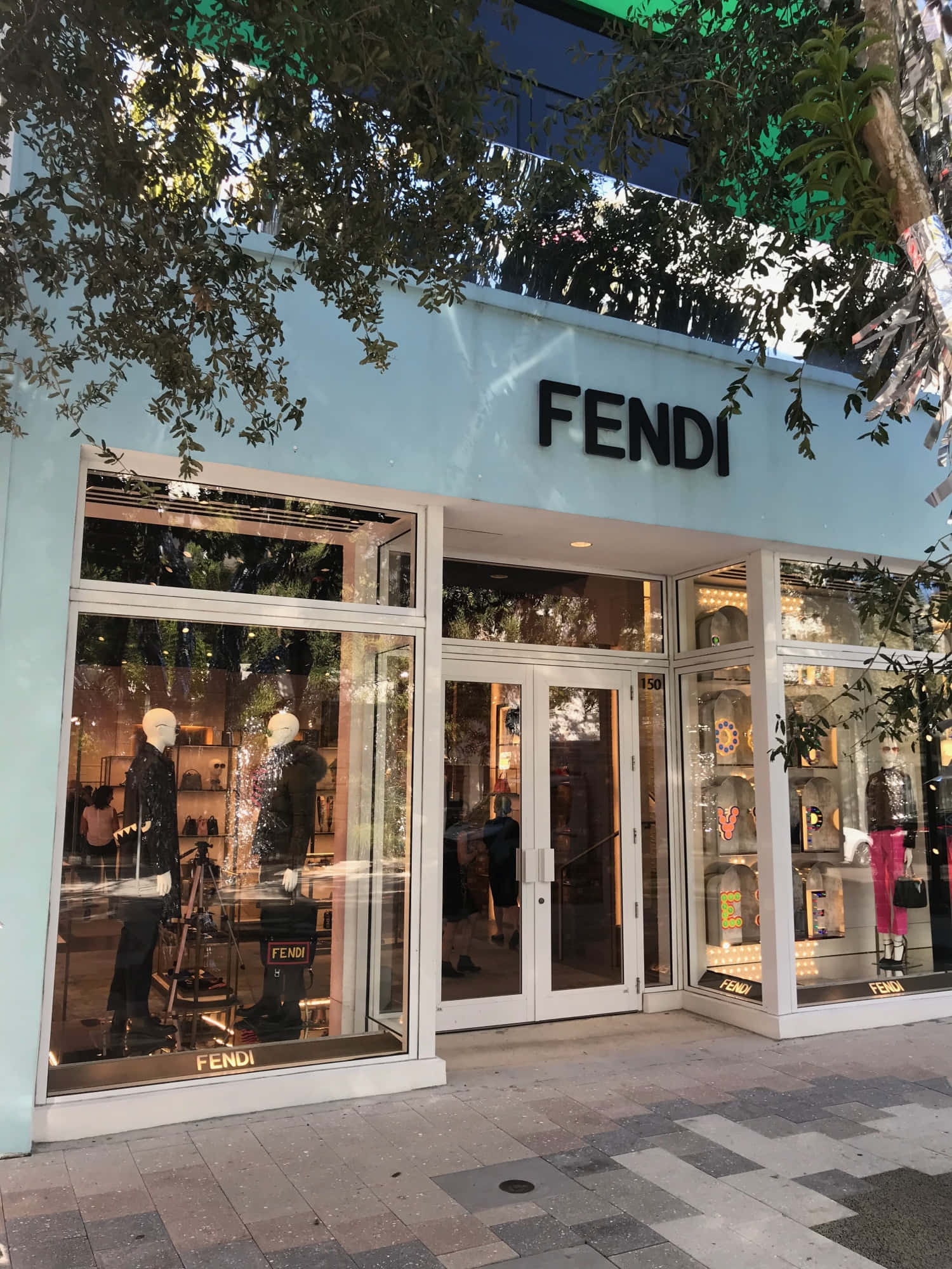 FENDI設計師手袋精選系列 壁紙 — 一間FENDI豪華精品店，淺藍色外牆和玻璃櫥窗，展示人體模型，上方樹枝投下斑駁陰影。