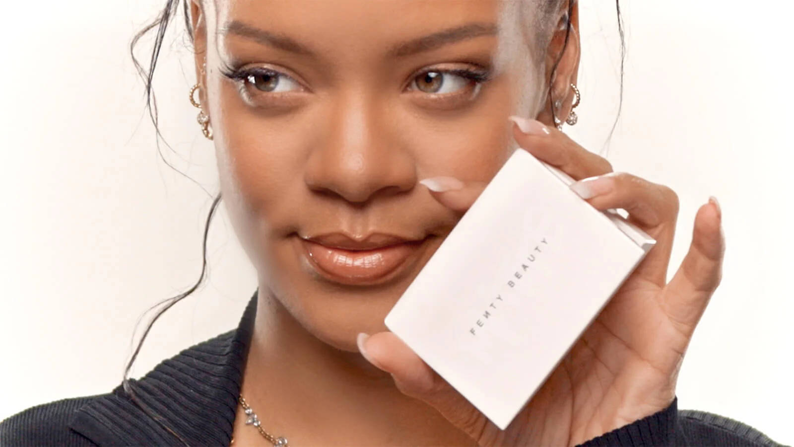 Rihanna 帶住溫柔嘅微笑，揸住一個 Fenty Beauty 化妝粉盒。