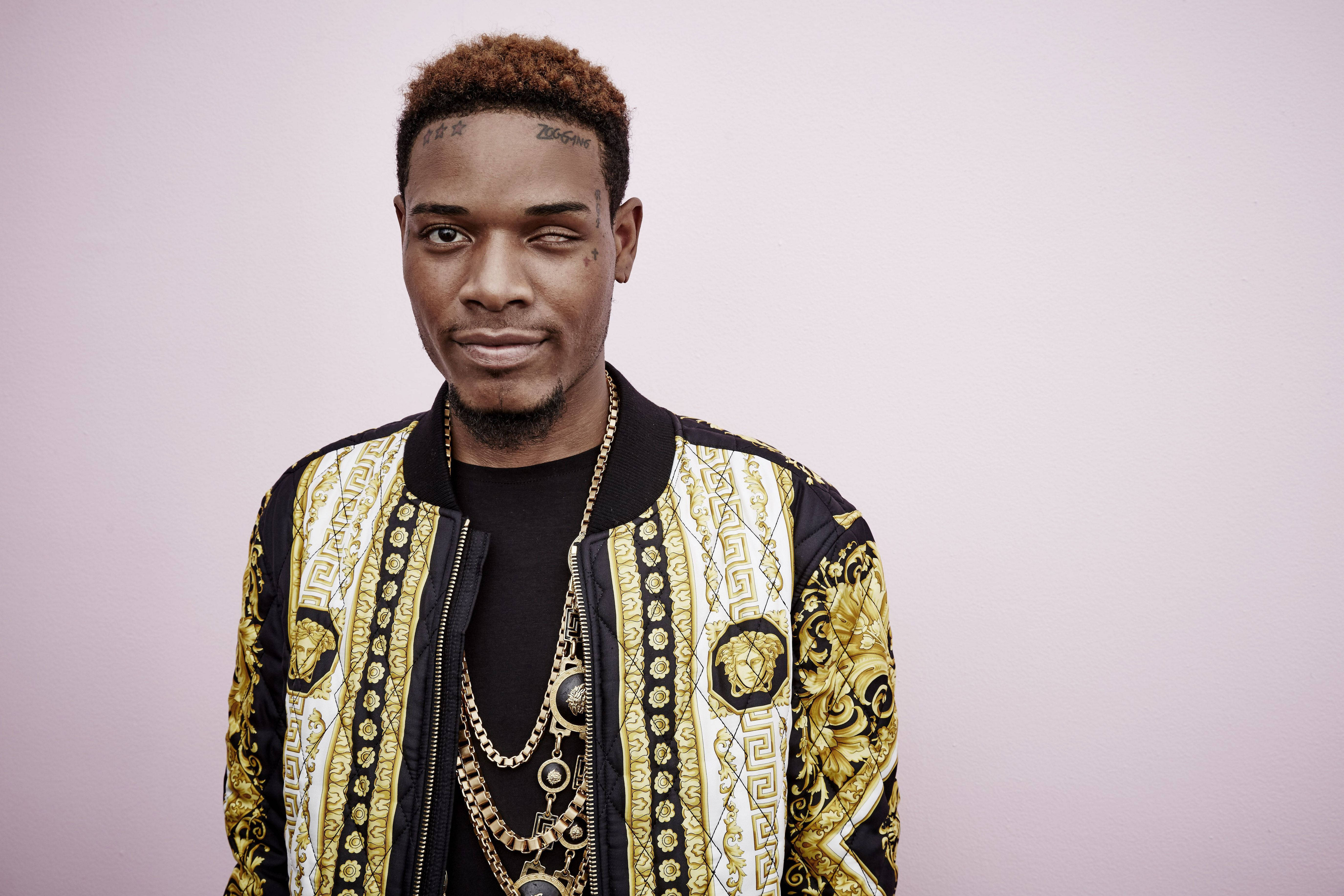 時尚Fetty Wap 壁紙 — 一位有紋身的時尚男士，在柔和的薰衣草背景前穿著金色和黑色圖案外套。