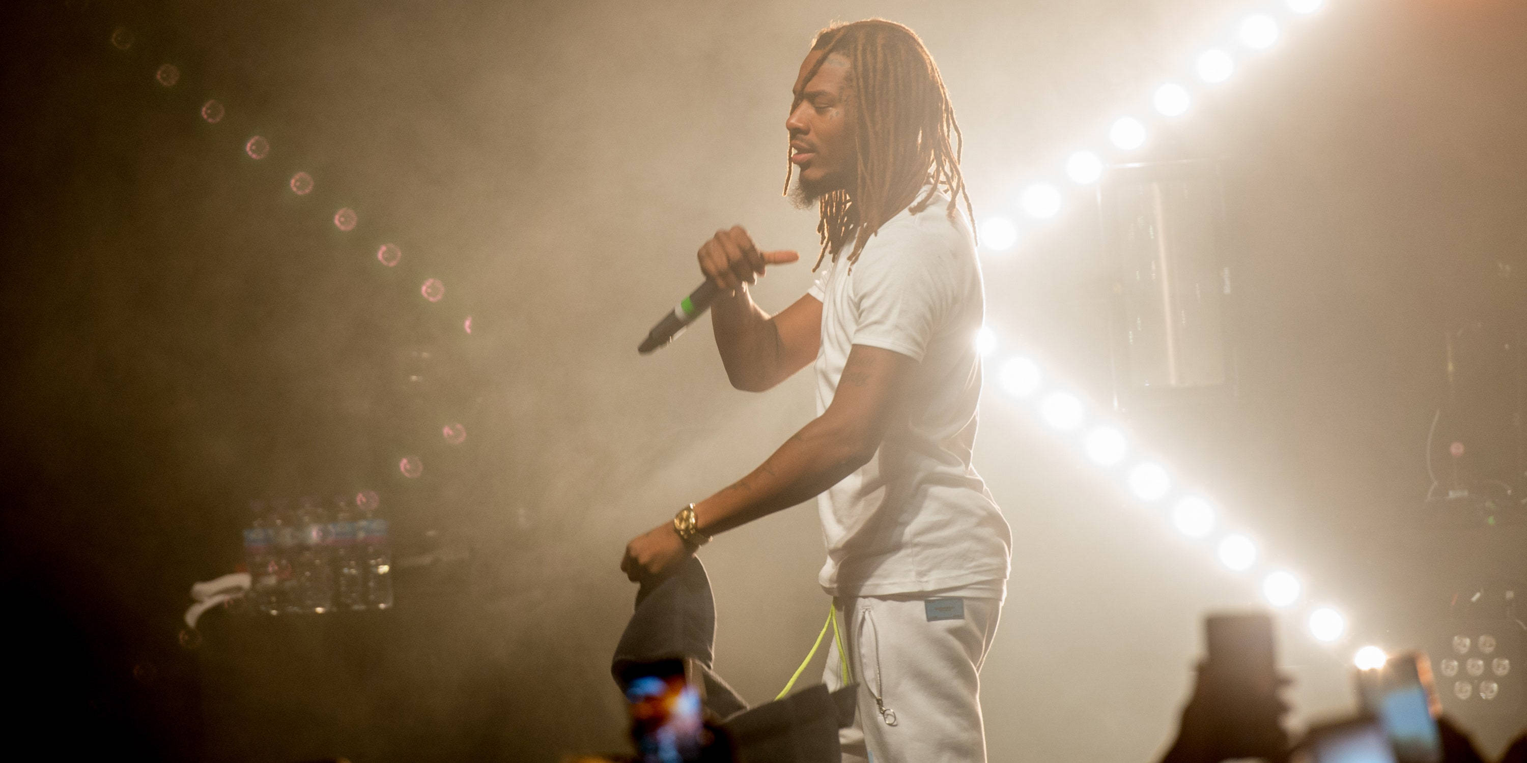 Fetty Wap 舞台燈光下 壁紙 — 一位留著辮子的男性表演者在明亮的舞台燈光下對著麥克風唱歌，煙霧和觀眾的手機清晰可見。