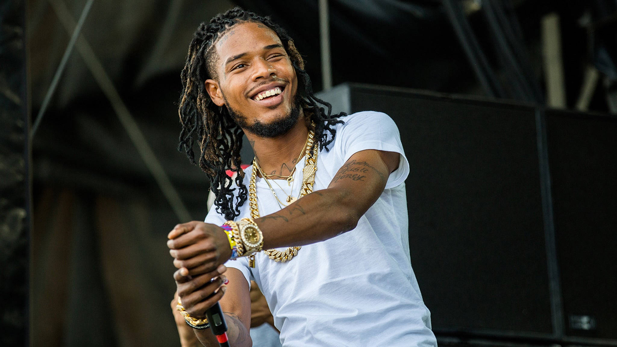 Fetty Wap 舞台表演 壁紙 — 一位微笑的饒舌歌手留著辮子頭，戴著金鏈，身穿白色T恤，手持麥克風在舞台上表演。