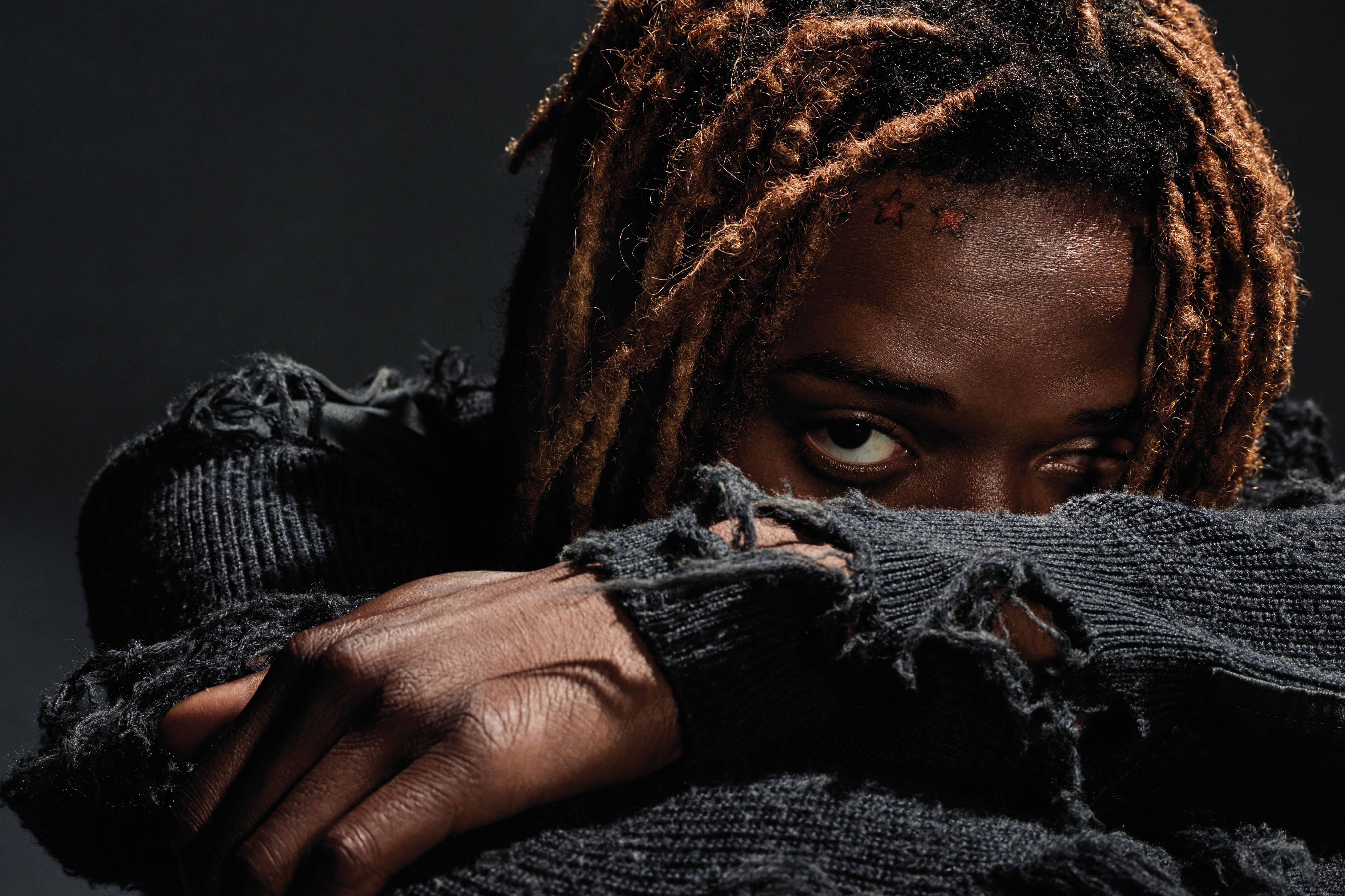 Fetty Wap 被手臂遮臉 壁紙 — 一個辮子頭髮和星星紋身的人，在黑暗背景中從一件破爛的黑色毛衣後面強烈地凝視。