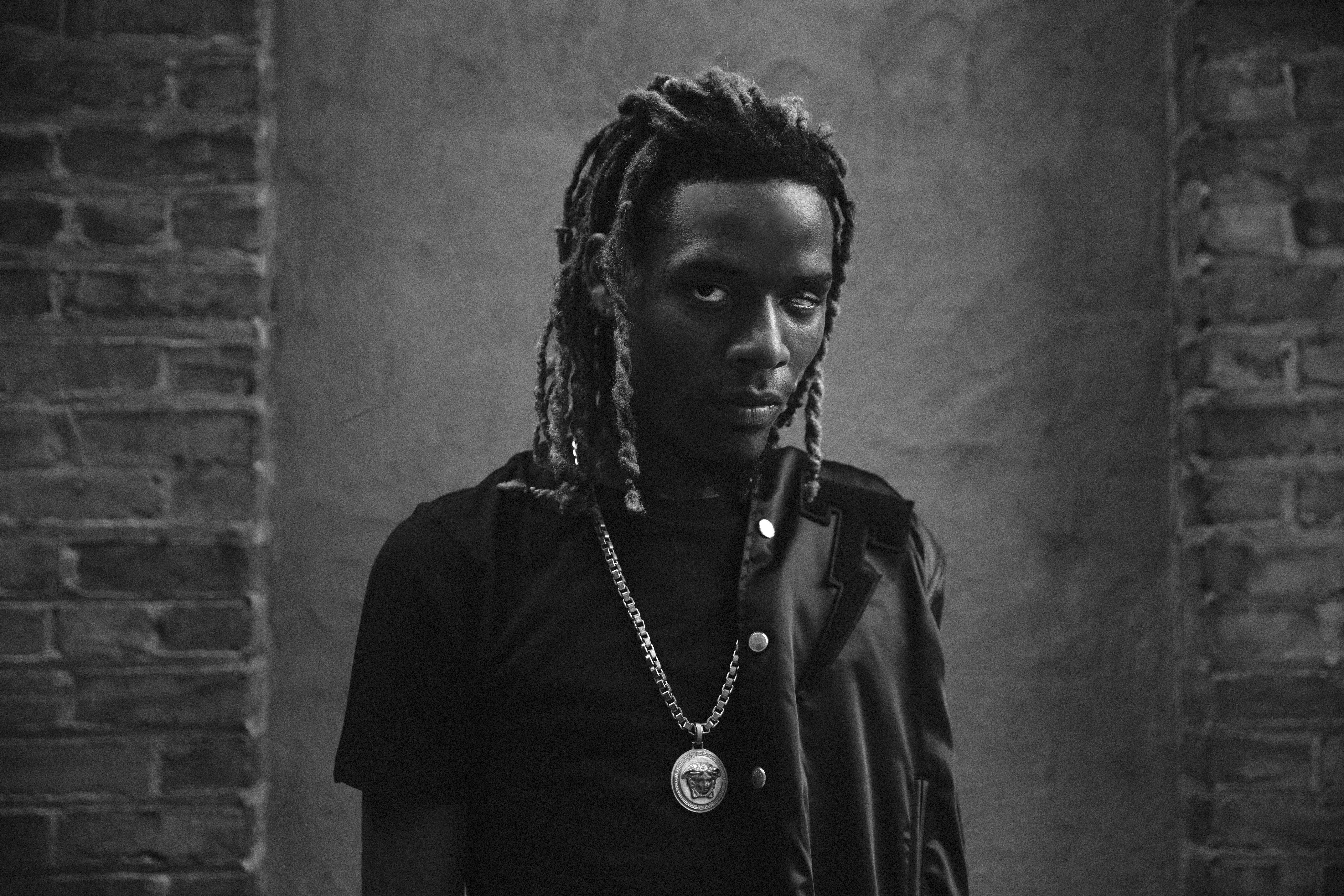 Fetty Wap 黑白攝影 壁紙 — 一個黑白肖像，一名留著辮子、身穿皮褸和戴著鏈條頸鏈的男子，背景是磚牆。