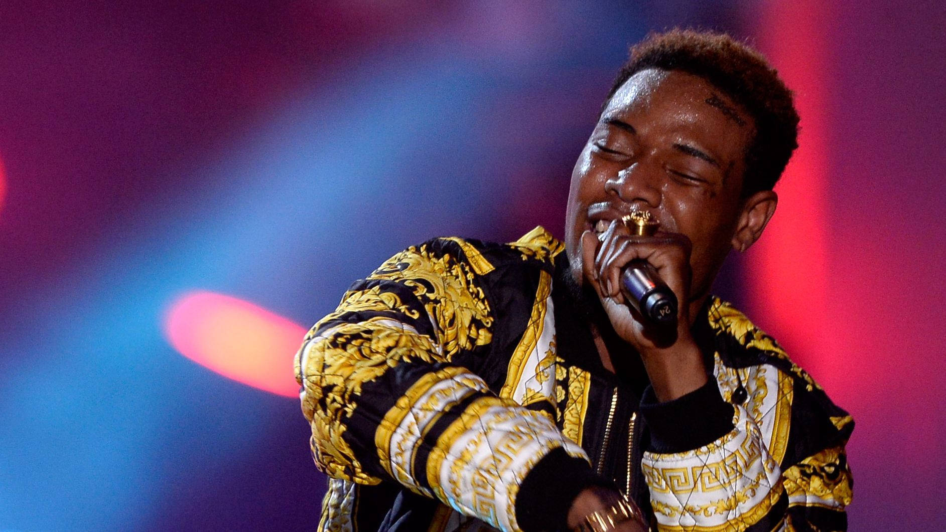 Fetty Wap演唱 壁紙 — 一位身穿金色和黑色圖案外套的男性表演者在充滿活力的舞台燈光下熱情地對著麥克風唱歌。