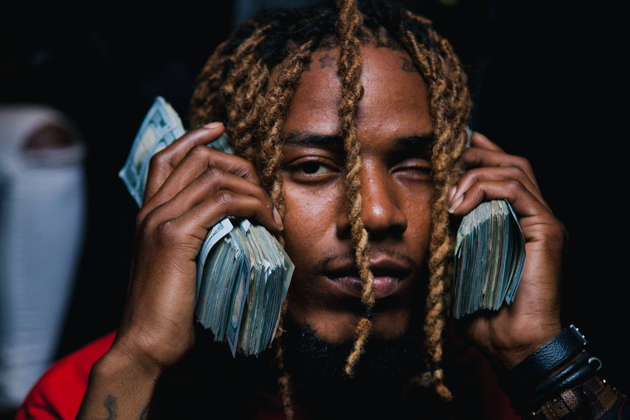 Fetty Wap特寫手持現金 壁紙 — 一個梳辮子的男人在深色背景前，像戴耳機一樣將一疊疊鈔票貼在耳邊。