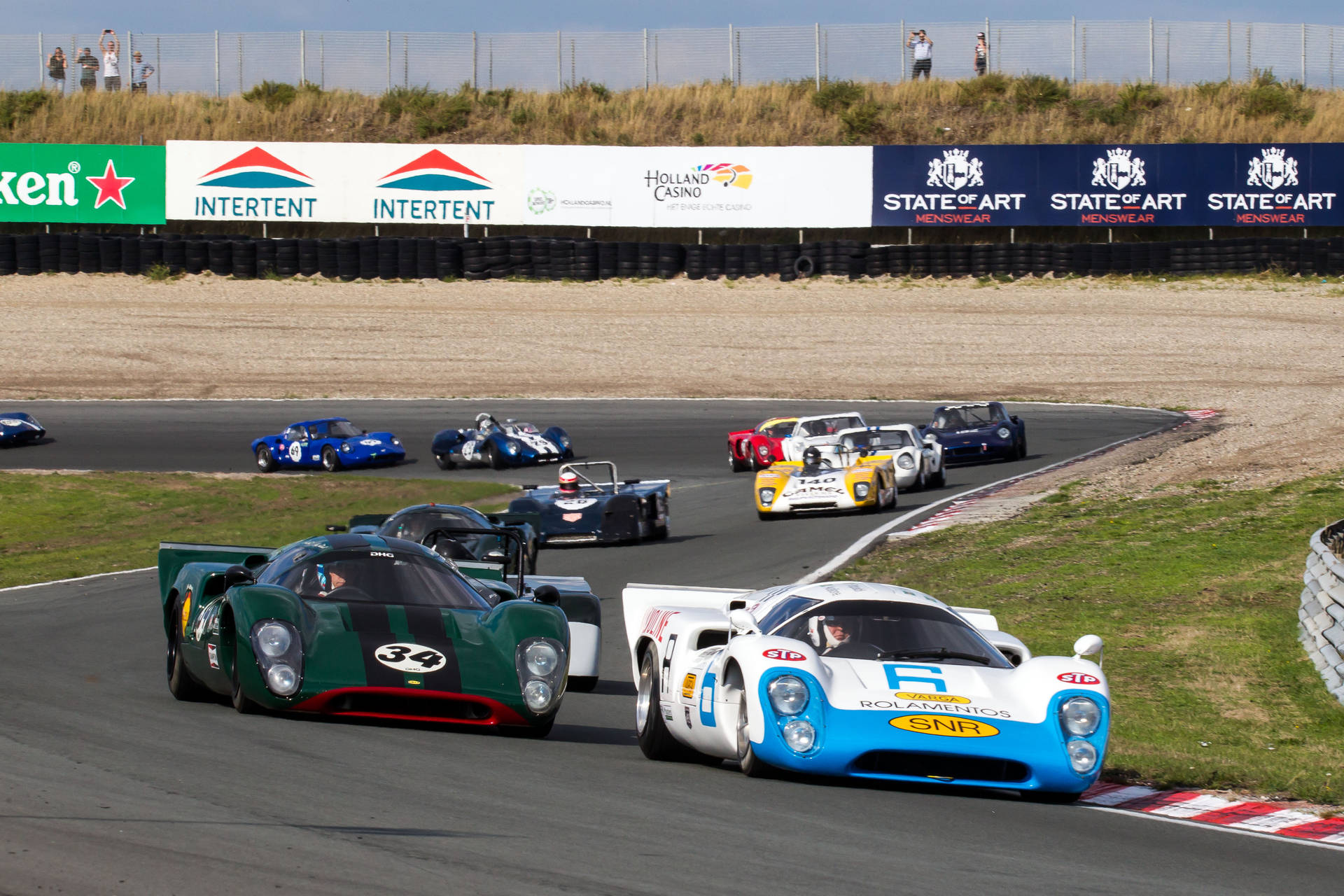 FIA Masters 歷史賽車錦標賽 壁紙 — 經典賽車在陽光普照的賽道上呼嘯而過，觀眾在草坡上觀看。