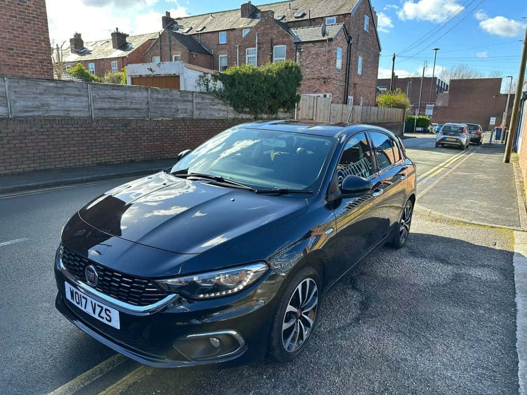 Fiat Tipo 桌布 壁紙 — 一架型格黑色Fiat Punto停泊在陽光明媚的住宅街道上，背景有磚屋和木柵欄。