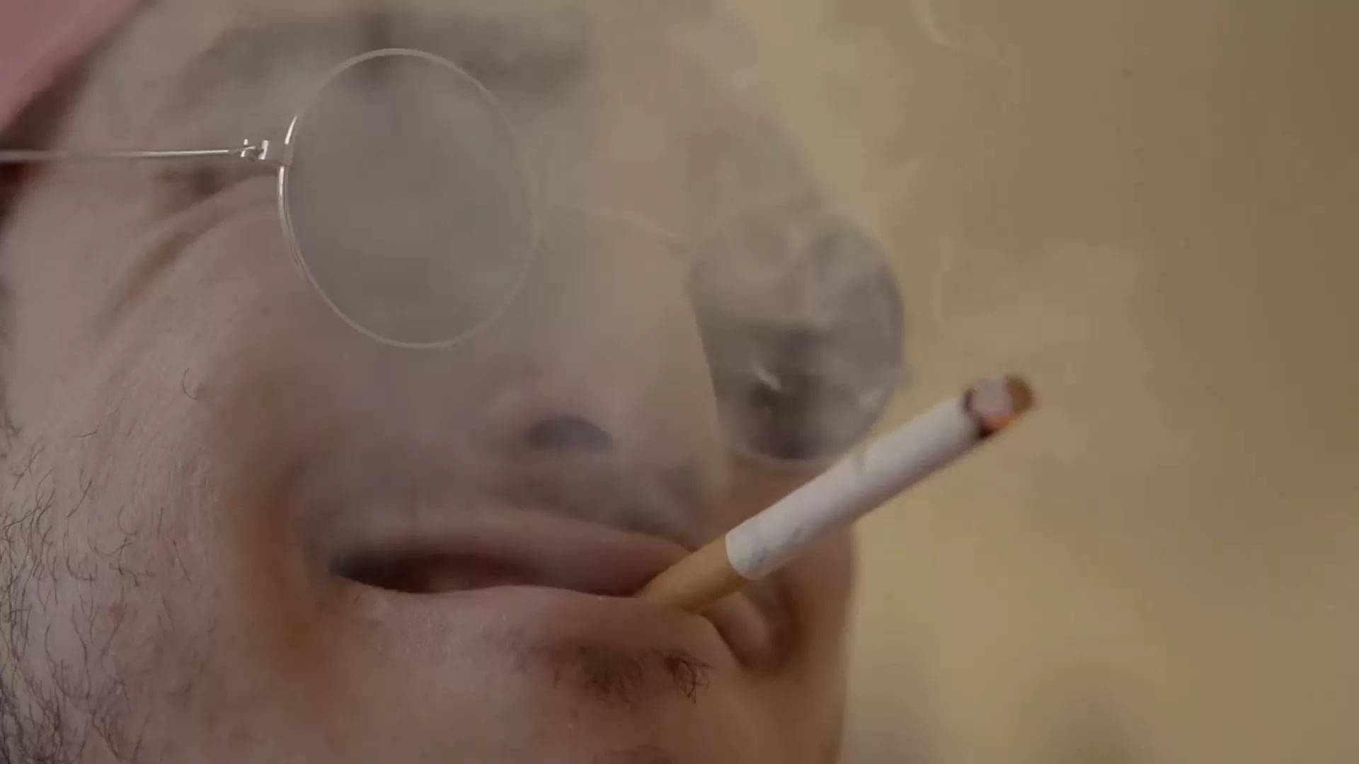 戴圓框眼鏡的男子呼出煙霧，手持香煙。