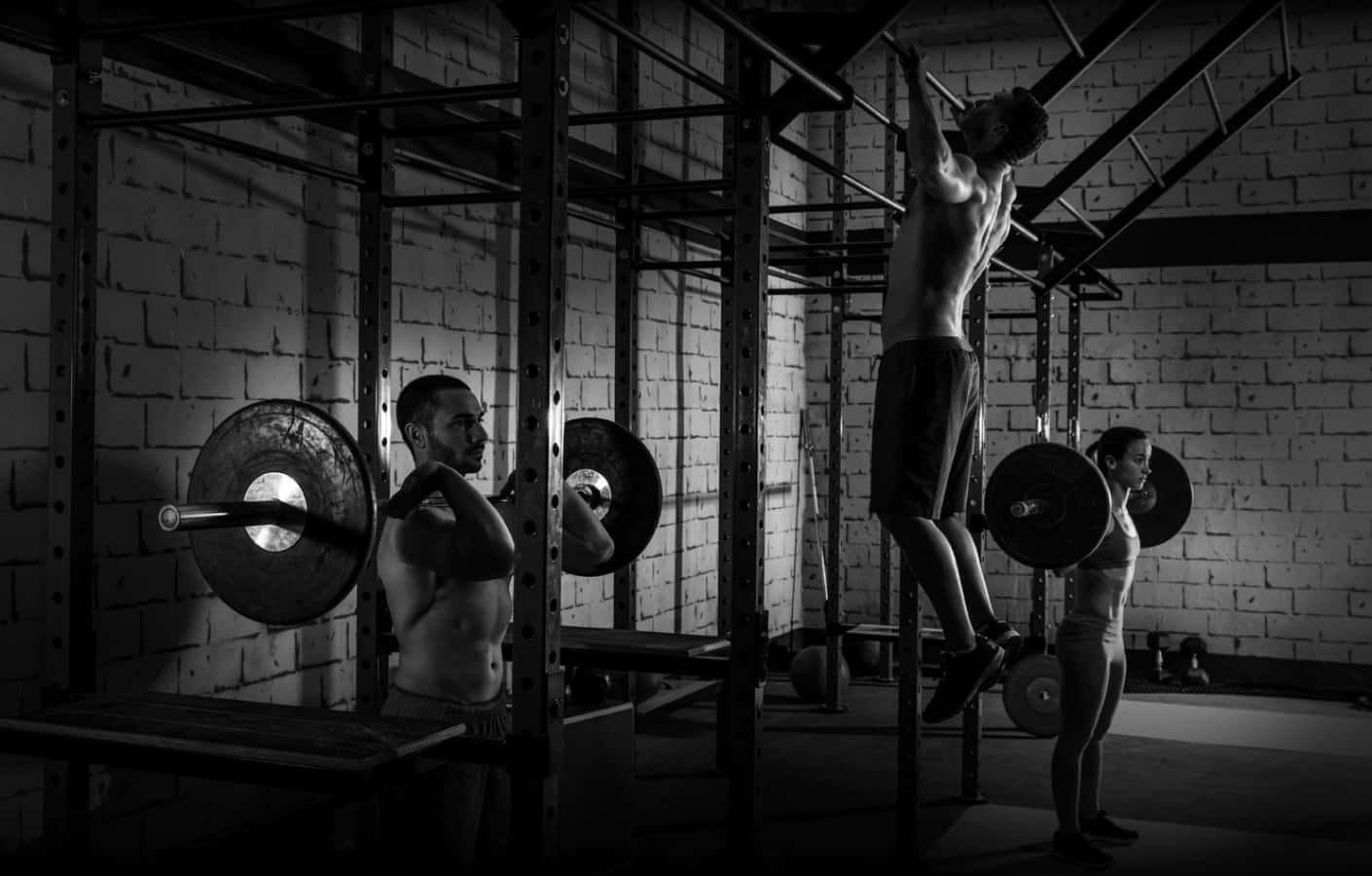 三位赤裸上身的運動員在光線昏暗的磚牆健身房中舉重。
