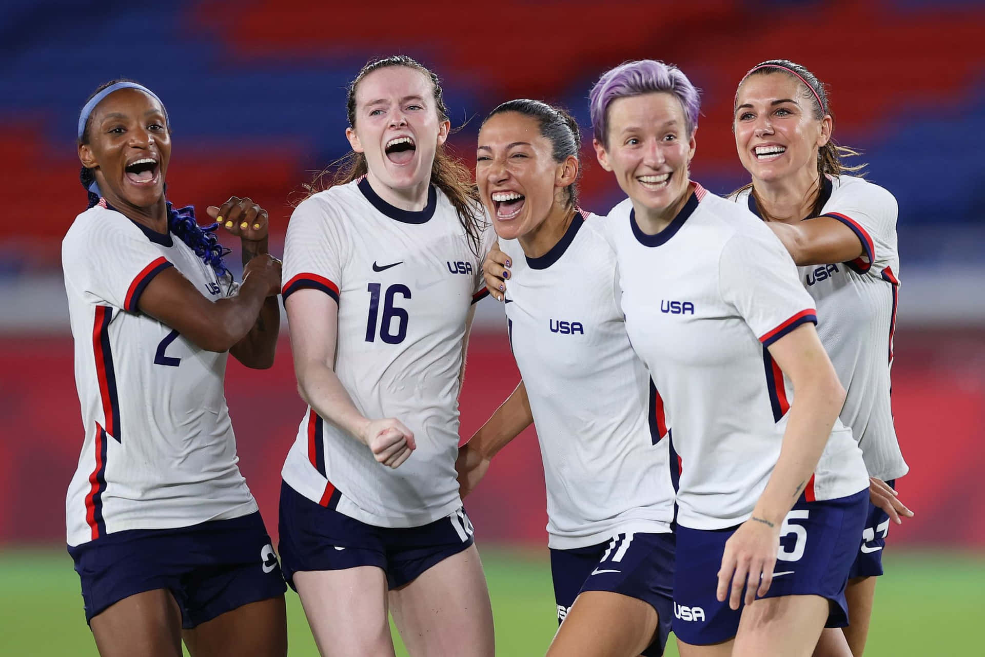  壁紙 — 五名身穿白色球衣、歡欣鼓舞的美國女子足球運動員在體育場上以燦爛的笑容和舉起的拳頭慶祝入球。