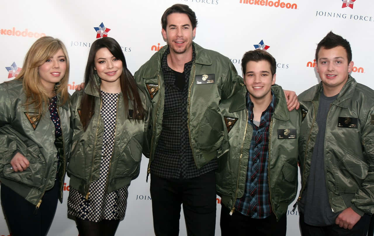  壁紙 — 五個微笑嘅人穿着相配嘅橄欖綠色外套，喺一個 Nickelodeon 活動中，喺有品牌標誌嘅背景前一齊擺姿勢。