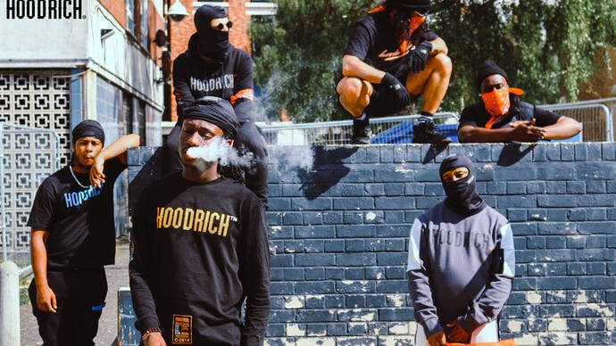 五名身穿Hoodrich街頭服的年輕男性在藍色磚牆上與煙霧擺姿勢。