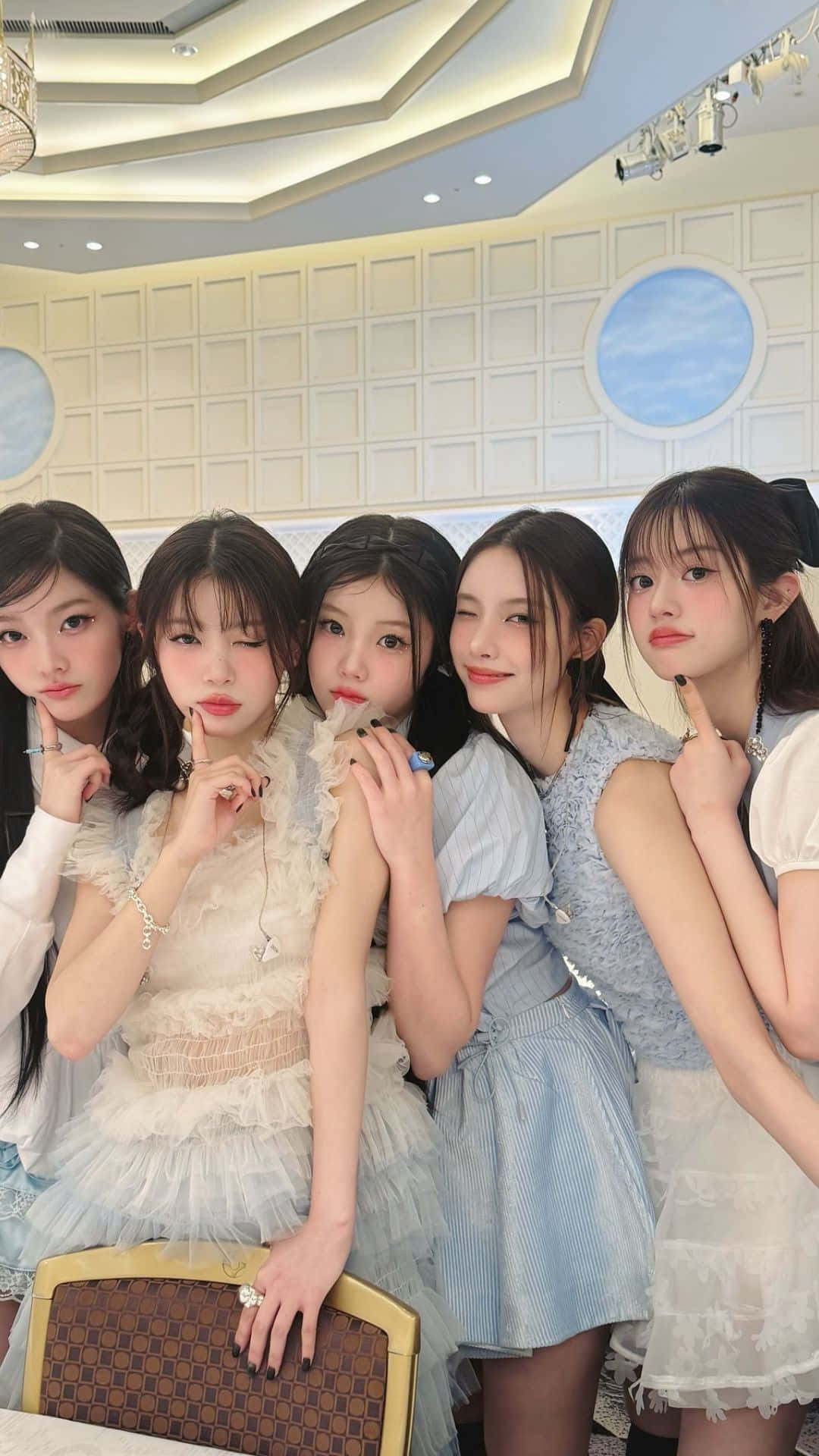  壁紙 — 五位年輕女性身穿粉彩連衣裙，在明亮的吊燈下以手指輕觸嘴唇的姿勢在室內俏皮地擺姿勢。