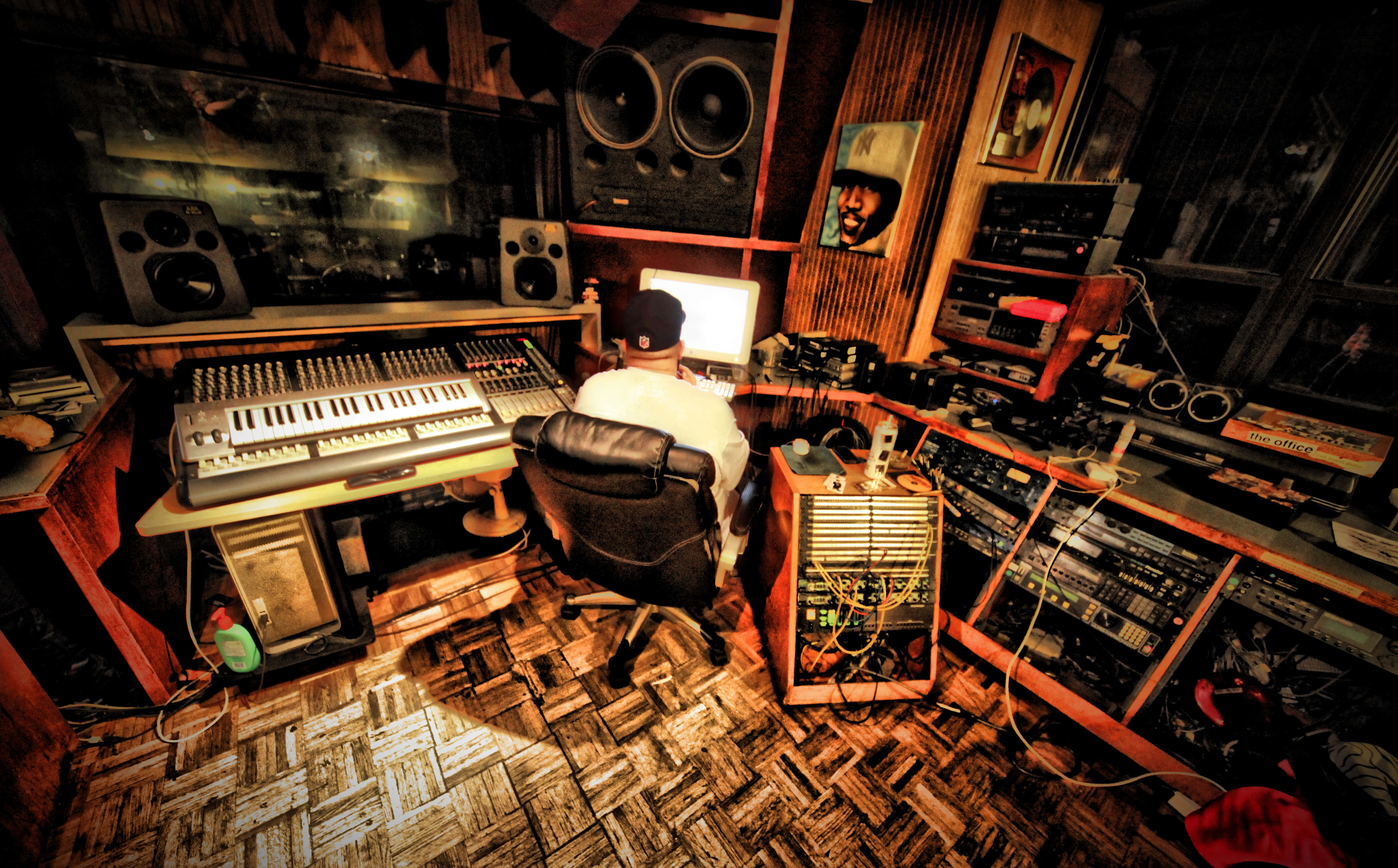 FL Studio 內的音樂器材 壁紙 — 一位音樂家坐在雜亂的工作室桌前，周圍環繞著復古音響設備和發光的屏幕，在溫暖的琥珀色燈光下。