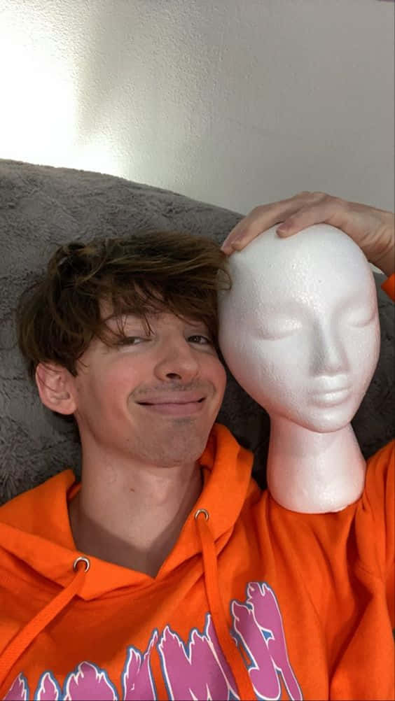 身穿橙色連帽衫的年輕男子在灰色沙發上的白色模特頭旁邊微笑。