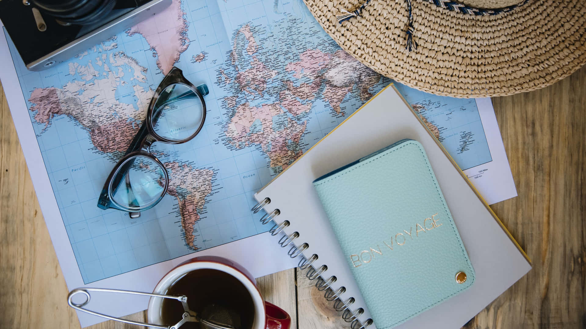  壁紙 — 一個平鋪的旅行裝備擺設，包括世界地圖、眼鏡、草帽和一本「Bon Voyage」筆記本，放在木質表面上。