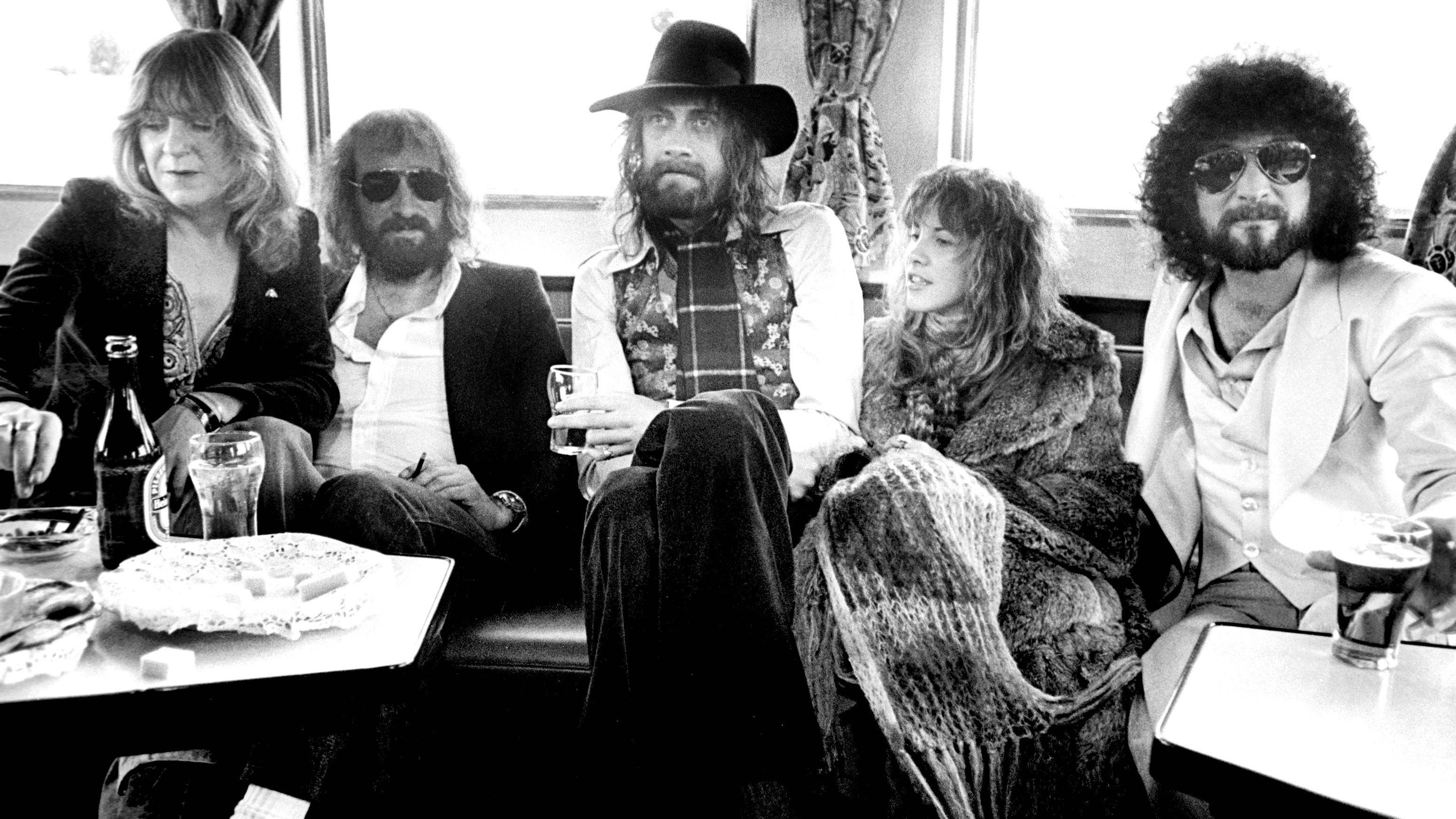 Fleetwood Mac 放鬆樂隊 壁紙 — 五位長髮音樂家穿著復古服裝，隨意地坐在火車長椅上，享用飲料和小吃。