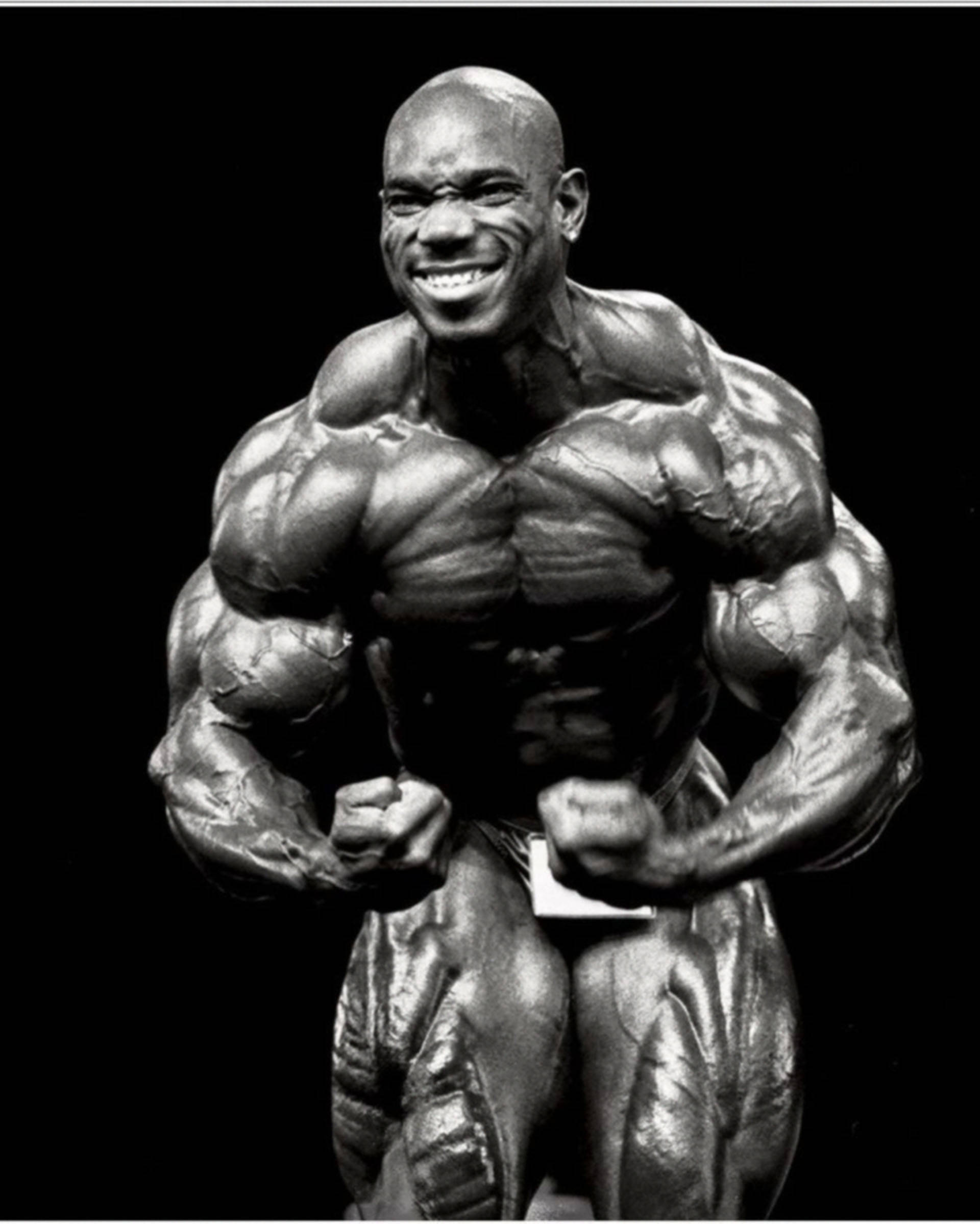運動型 Flex Wheeler 壁紙 — 一張黑白照片，顯示一位肌肉發達的健美運動員在深色背景前展露肌肉，露出燦爛的笑容。