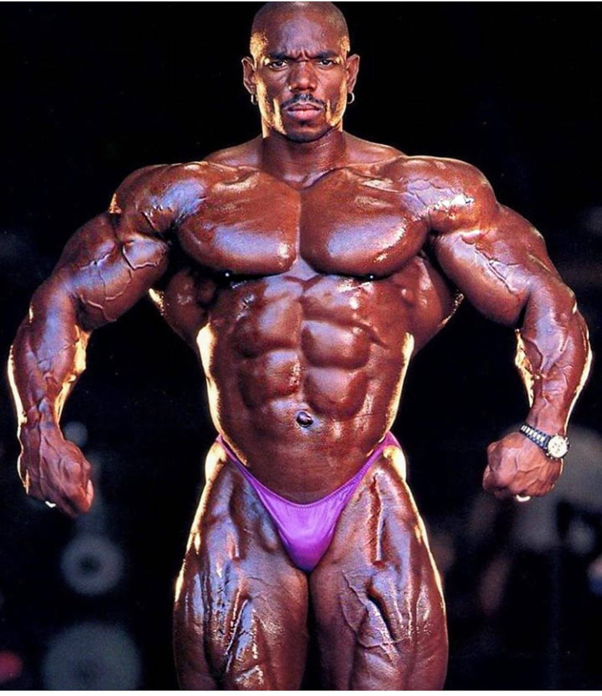 Flex Wheeler巔峰體格 壁紙 — 一位肌肉發達的健美運動員，皮膚塗油，在深色背景下穿著紫色比賽褲，擺出強烈的姿勢。
