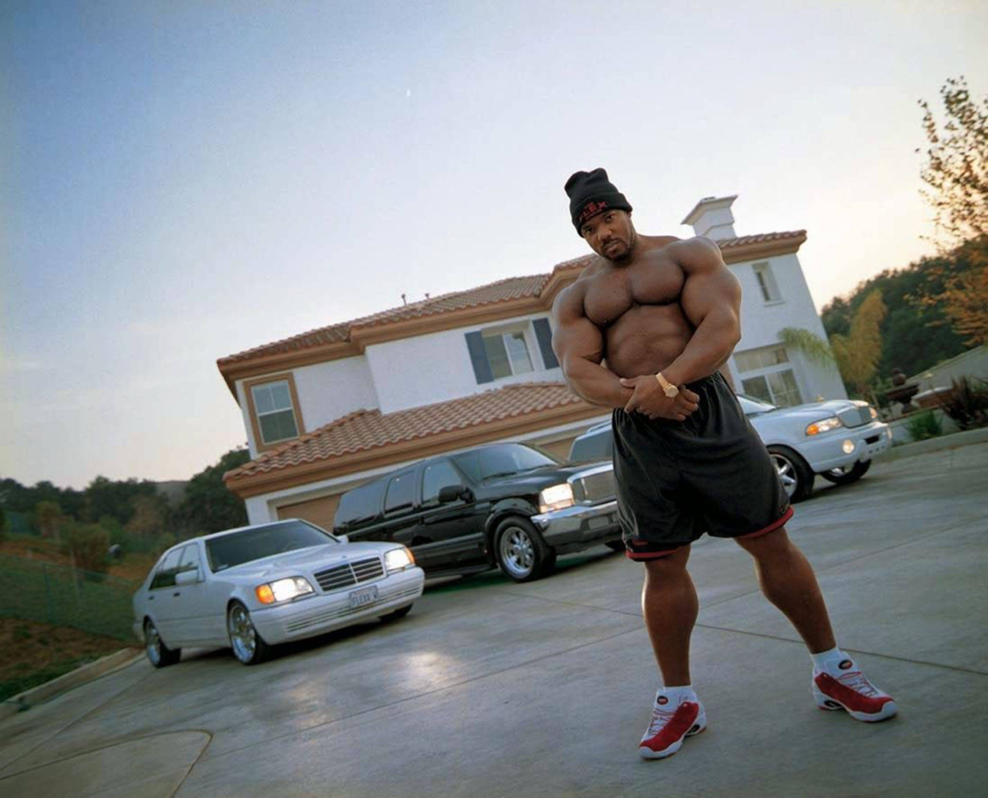 一個肌肉發達的男子在日落時分站在車道上，身後有豪華汽車和一棟房子。