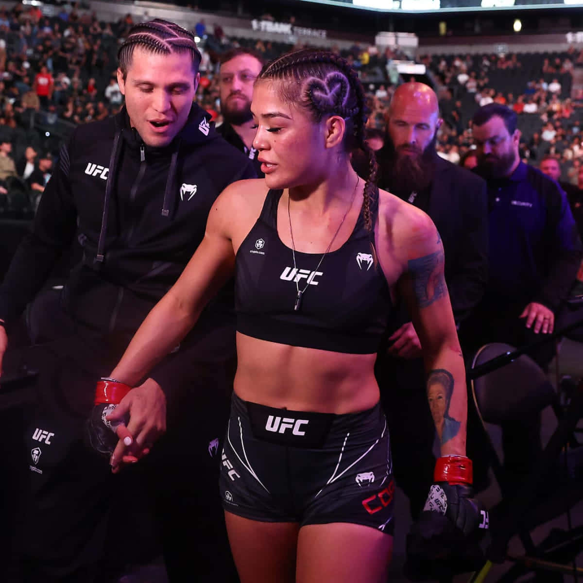  壁紙 — 一位專注的女性UFC拳手，梳著心形辮子，在場館燈光下握著教練的手走出八角籠。