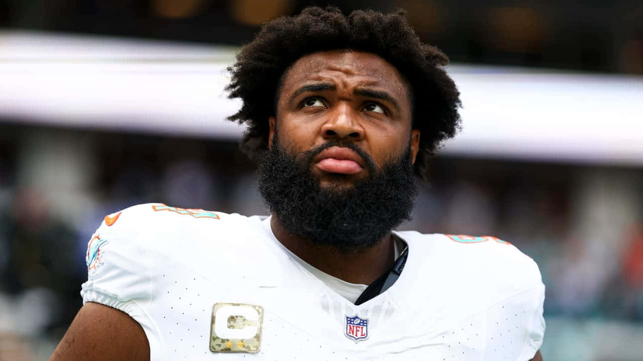  壁紙 — 一位留著濃密鬍鬚和非洲式髮型、專注的NFL球員，穿著帶有隊長臂章的白上衣，向上凝視。