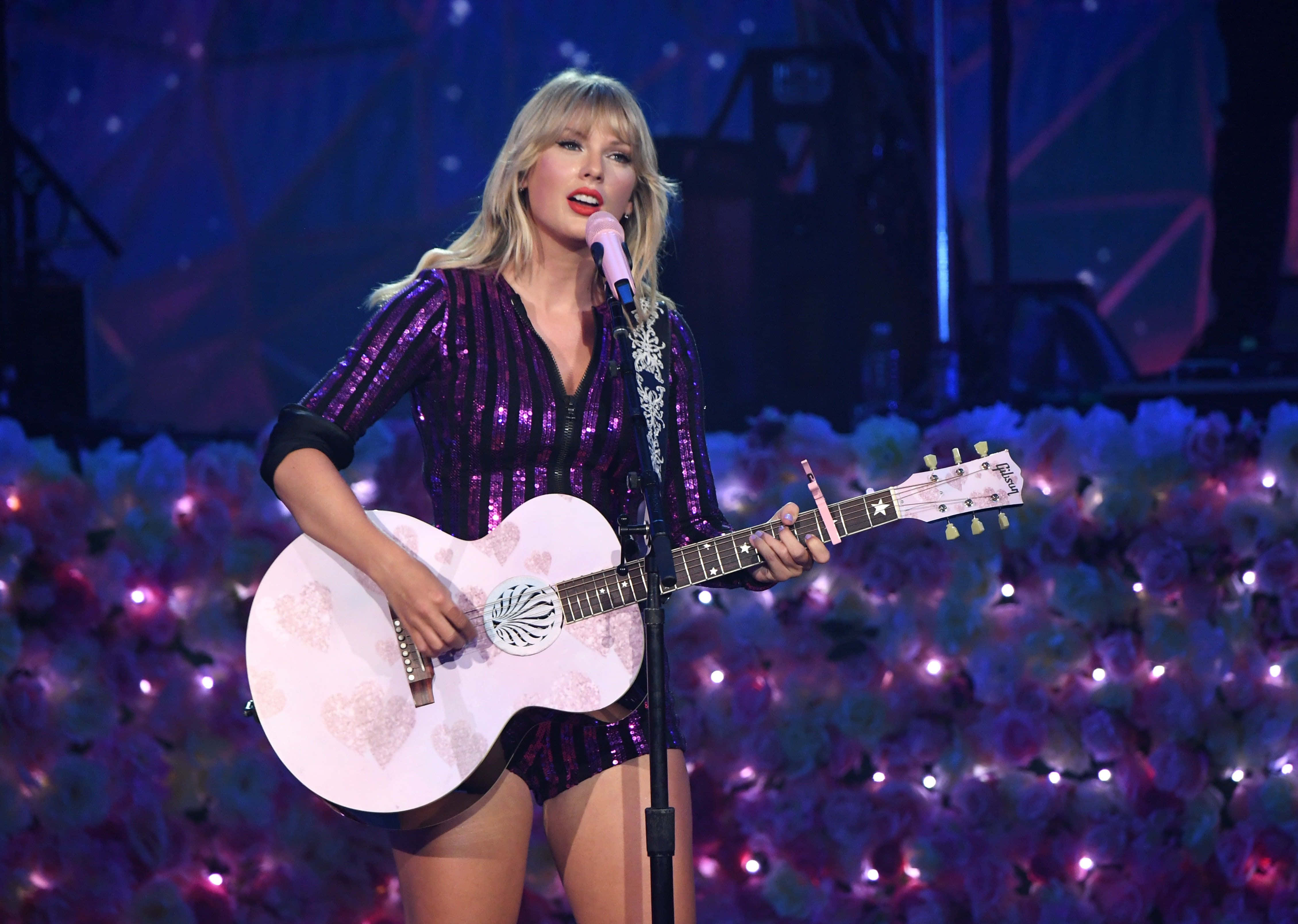 泰勒·斯威夫特為《Folklore》專輯展現迷人風采 壁紙 — Taylor Swift 在紫色舞台燈光下手持白色花卉木結他在舞台上表演。